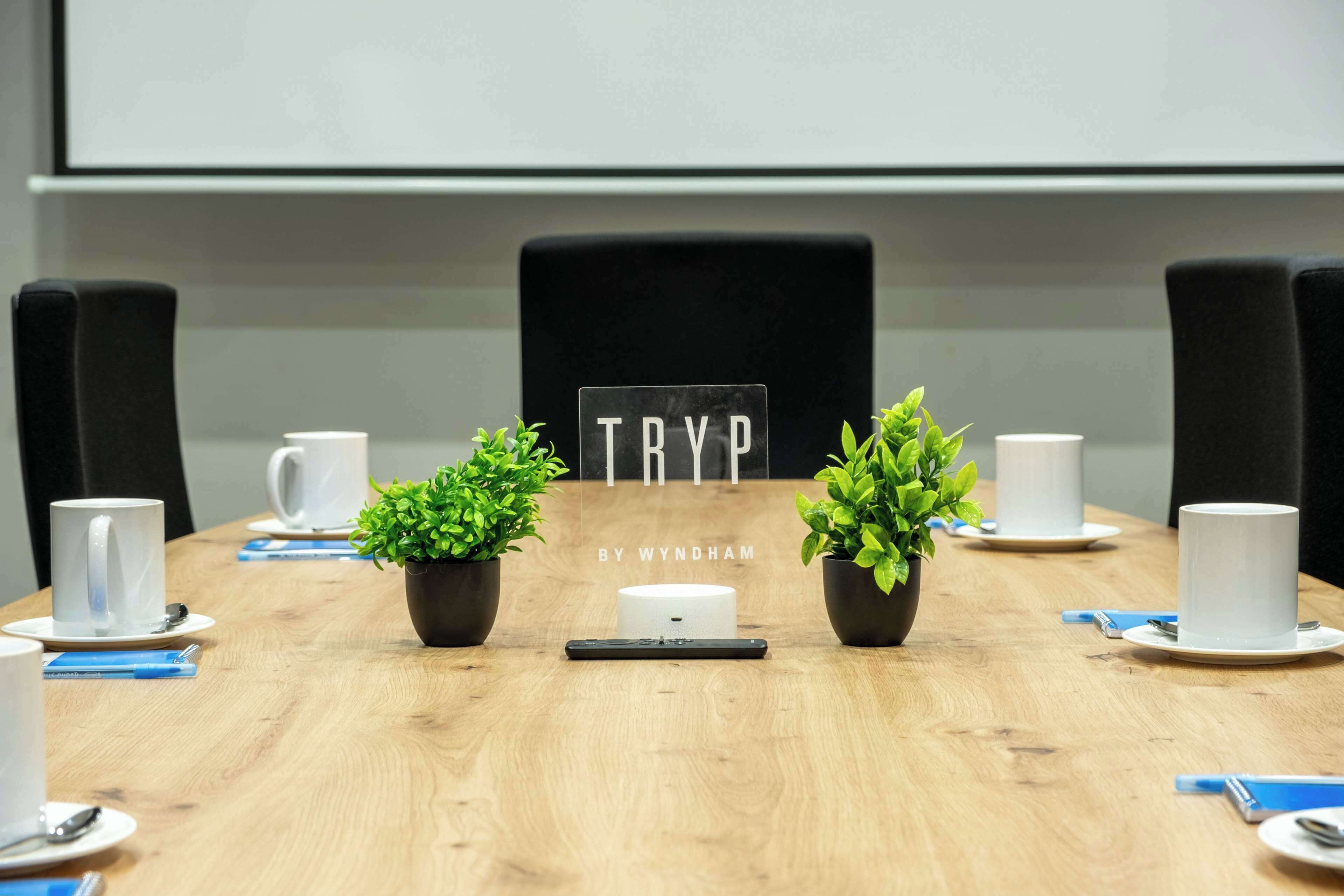 A conference room with a wooden table, white cups, black chairs, and small potted plants.