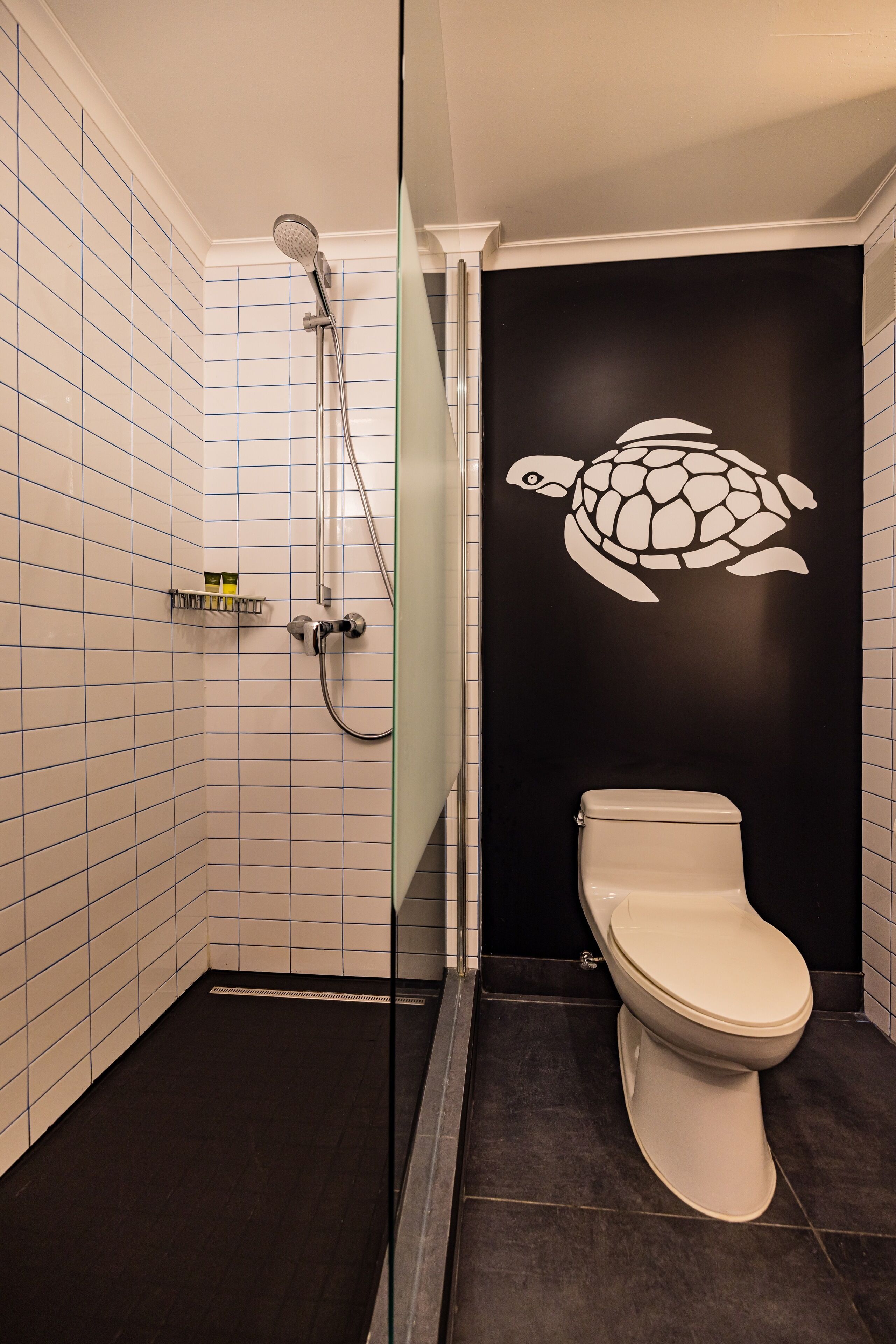 Un baño con ducha revestida de azulejos blancos, inodoro blanco y pared negra decorada con un vinilo de tortuga blanca.
