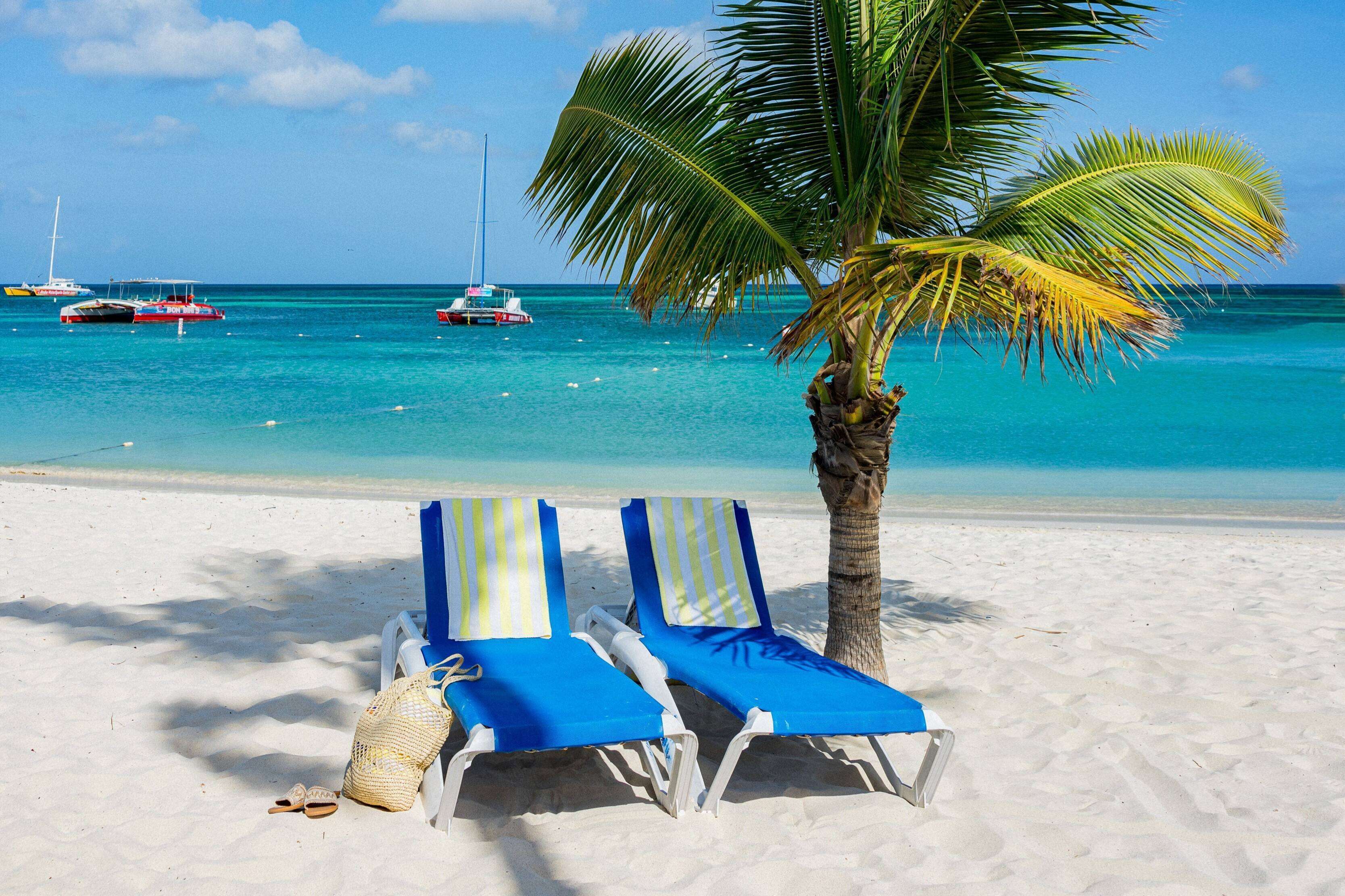 A beach with two lounge chairs, a palm tree, and boats in the water.