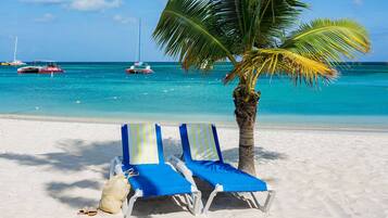 A beach with two lounge chairs, a palm tree, and boats in the water.