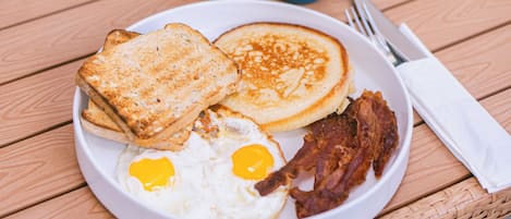 A breakfast plate with toast, pancakes, fried eggs, and bacon on a wooden table.