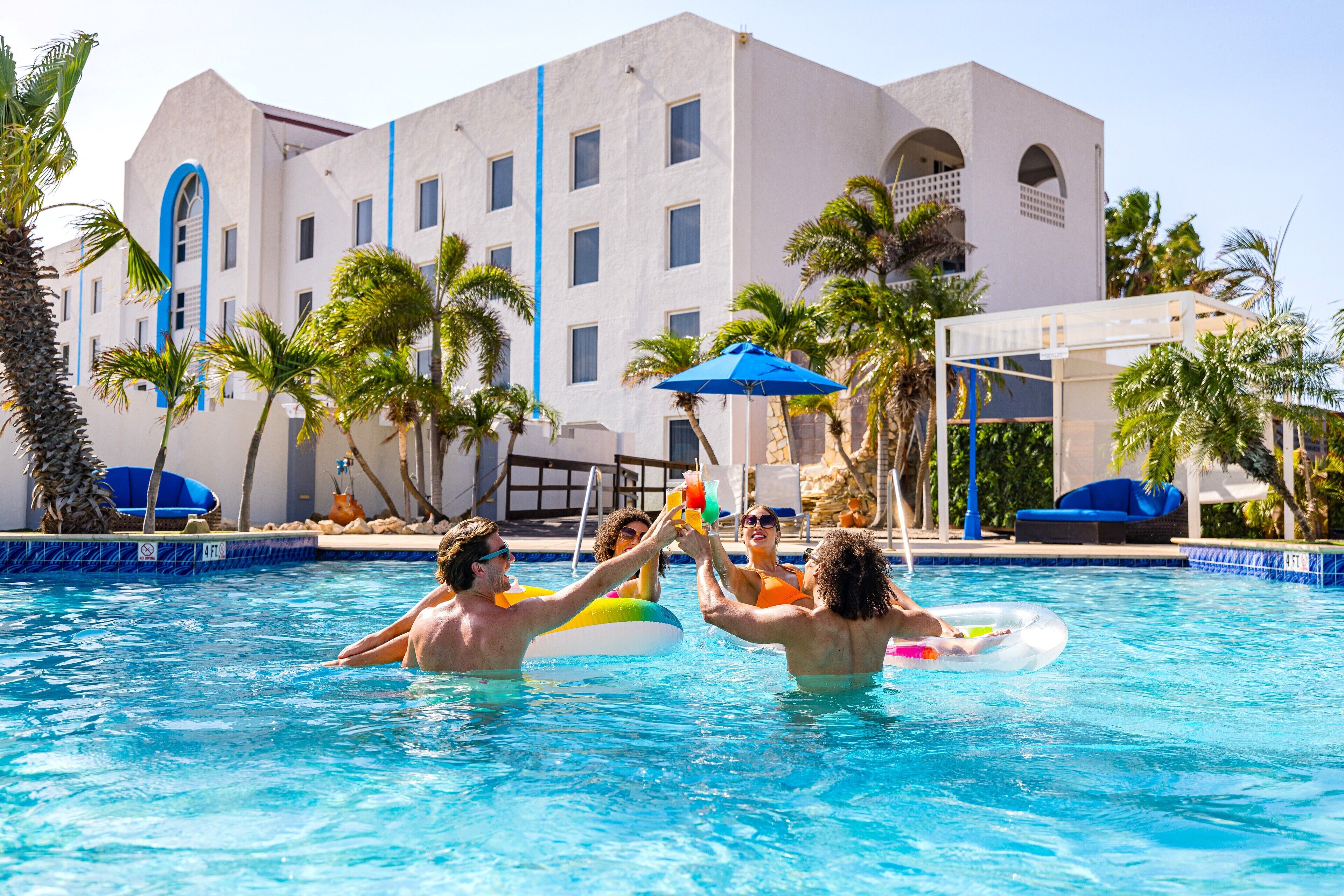 A swimming pool with people in the water, palm trees, and a building with blue accents in the background.