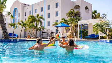 A swimming pool with people in the water, palm trees, and a building with blue accents in the background.