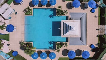 An aerial view of a pool area with a rectangular pool, surrounded by lounge chairs and umbrellas, and palm trees.