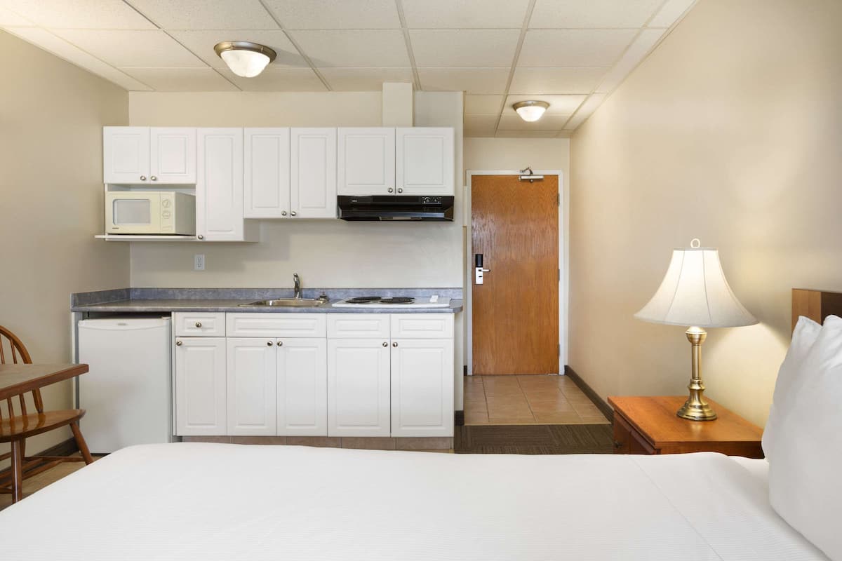A compact hotel room with a kitchenette, a bed, a bedside table with a lamp, and a wooden chair.
