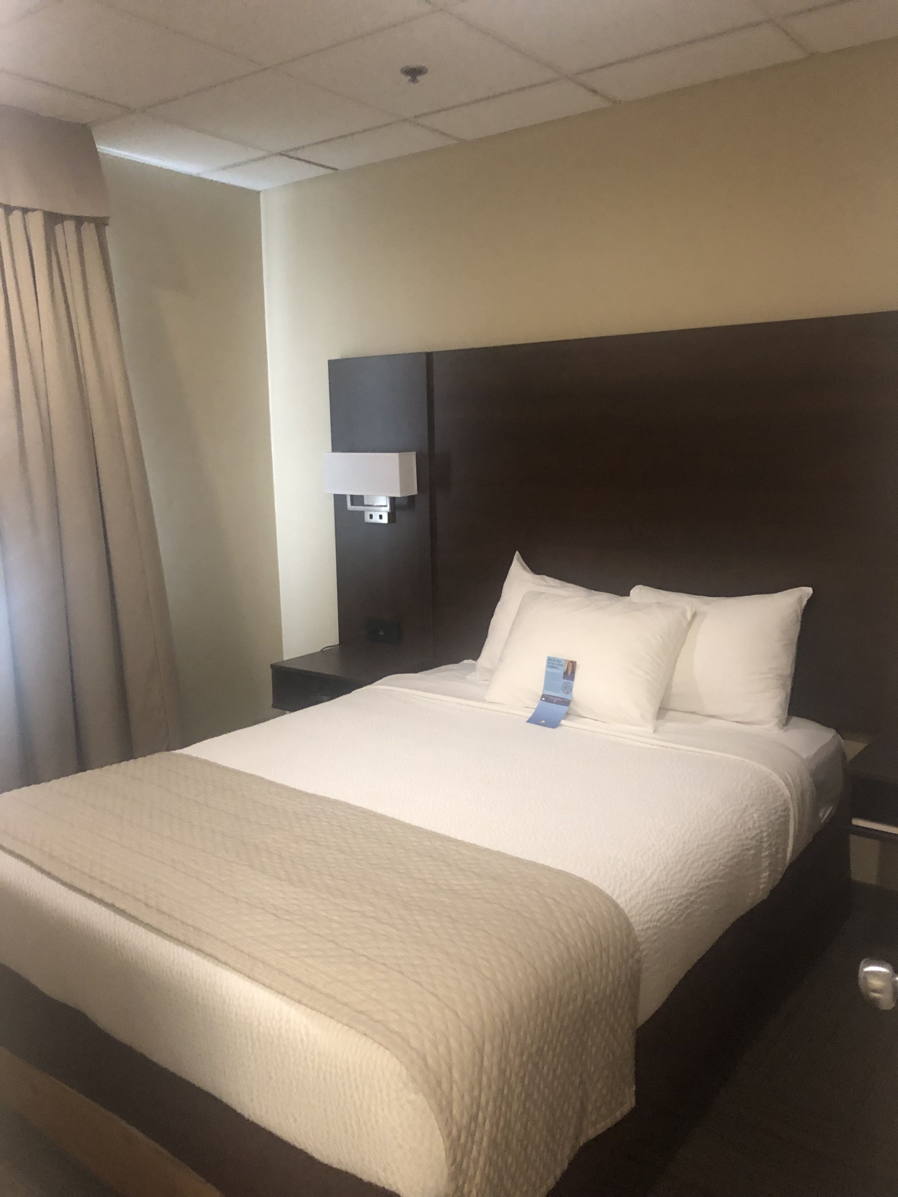 Habitación de hotel con una cama grande, una mesita de noche, una lámpara de pared y una ventana con cortinas.
