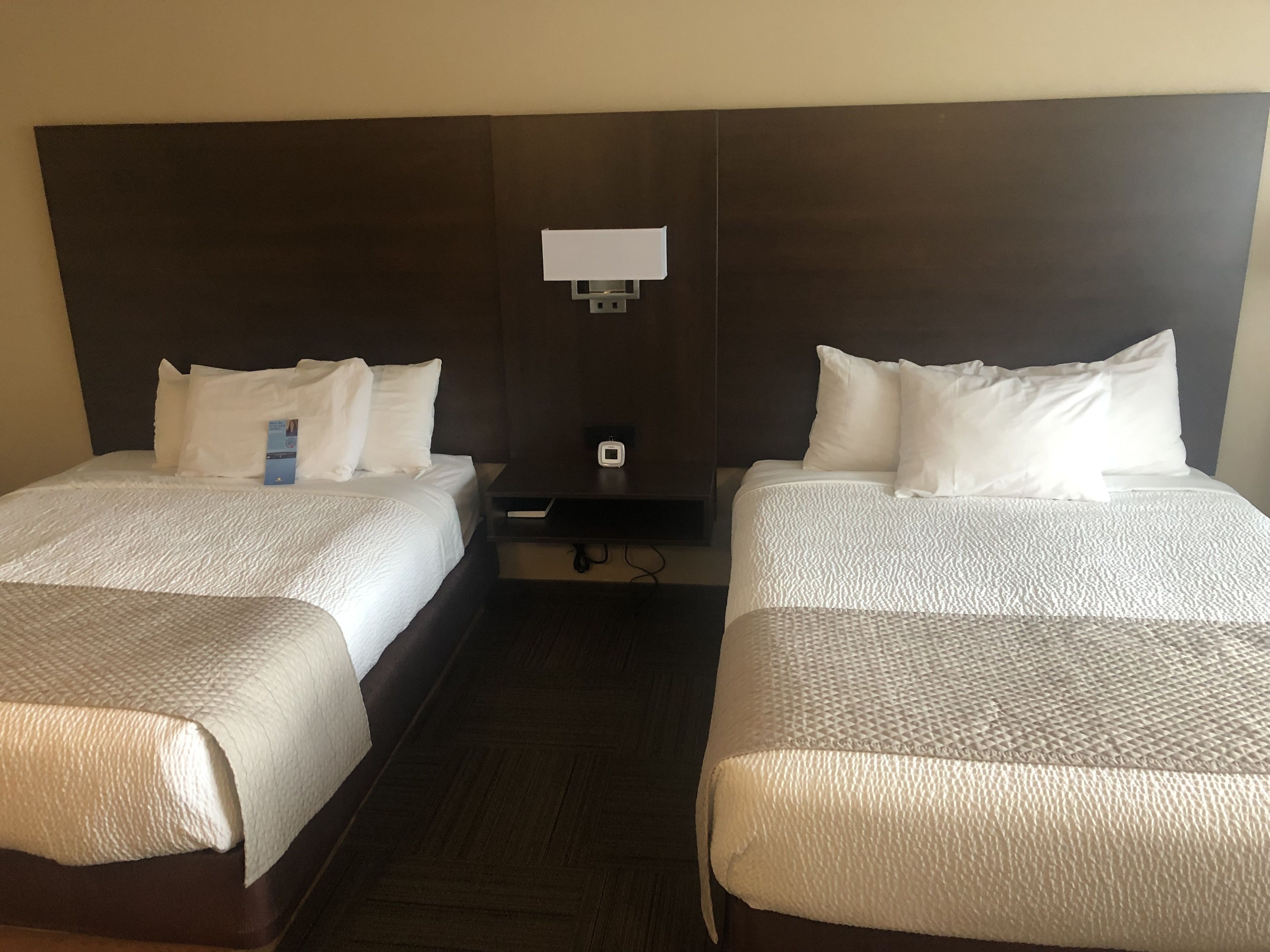 A hotel room with two beds, each with white bedding and pillows. There is a small table between the beds with a clock and a power outlet.