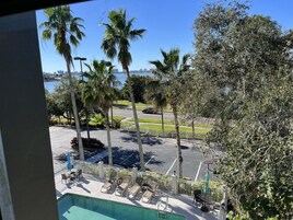 A poolside area with lounge chairs, a swimming pool, palm trees, and a view of the ocean and city skyline.