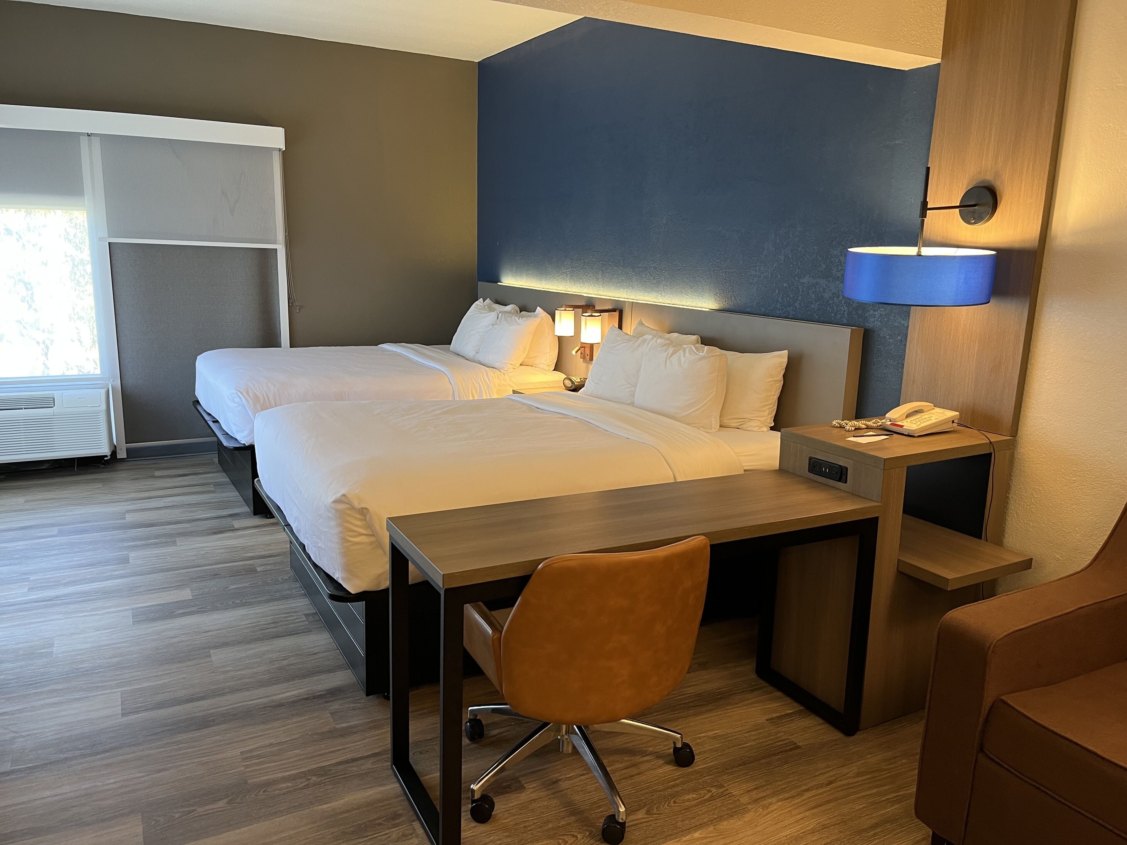 A hotel room with two beds, a desk, a chair, a phone, and a lamp.
