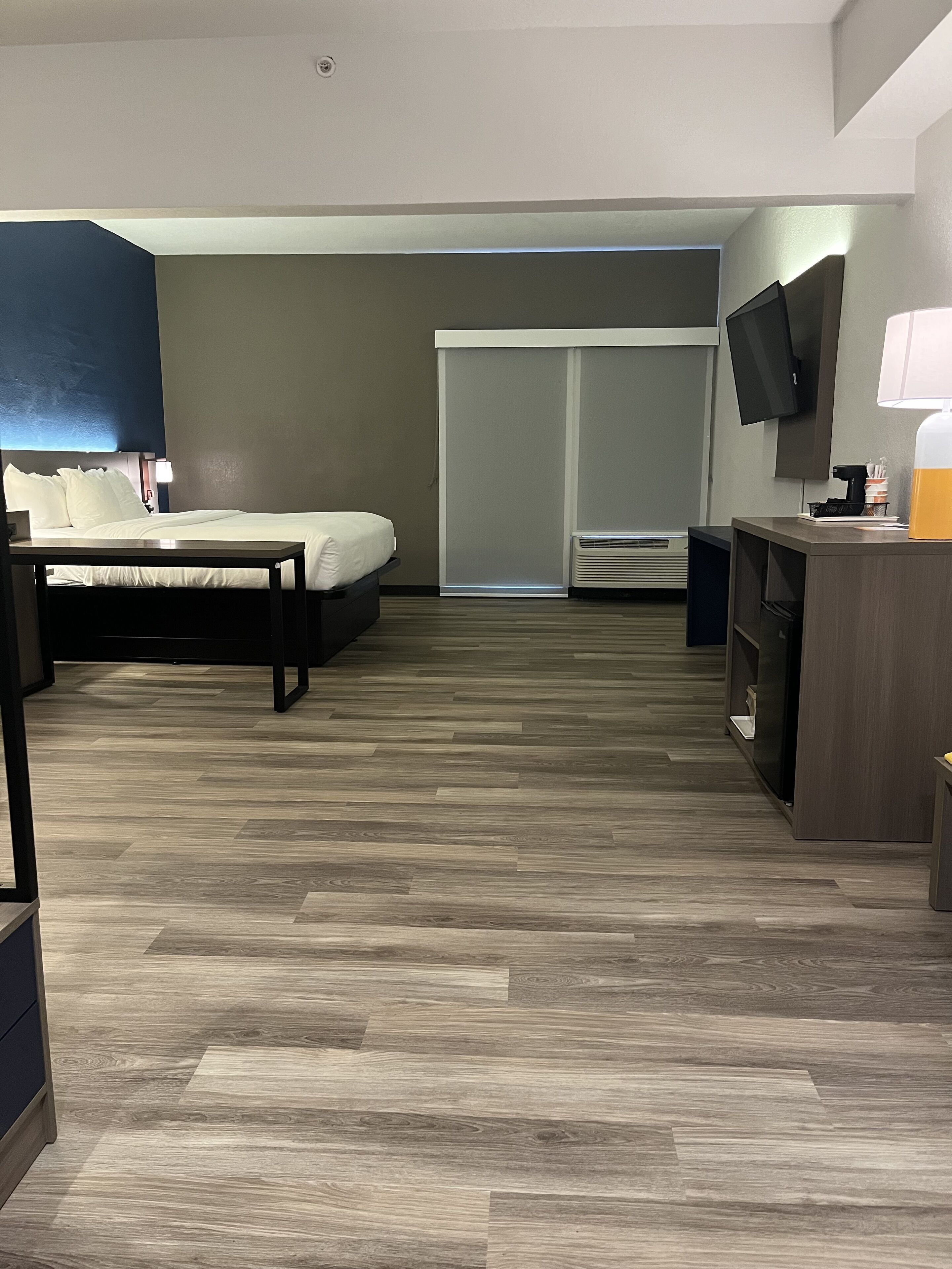 A hotel room with a bed, bedside table, TV, and a desk with a lamp.