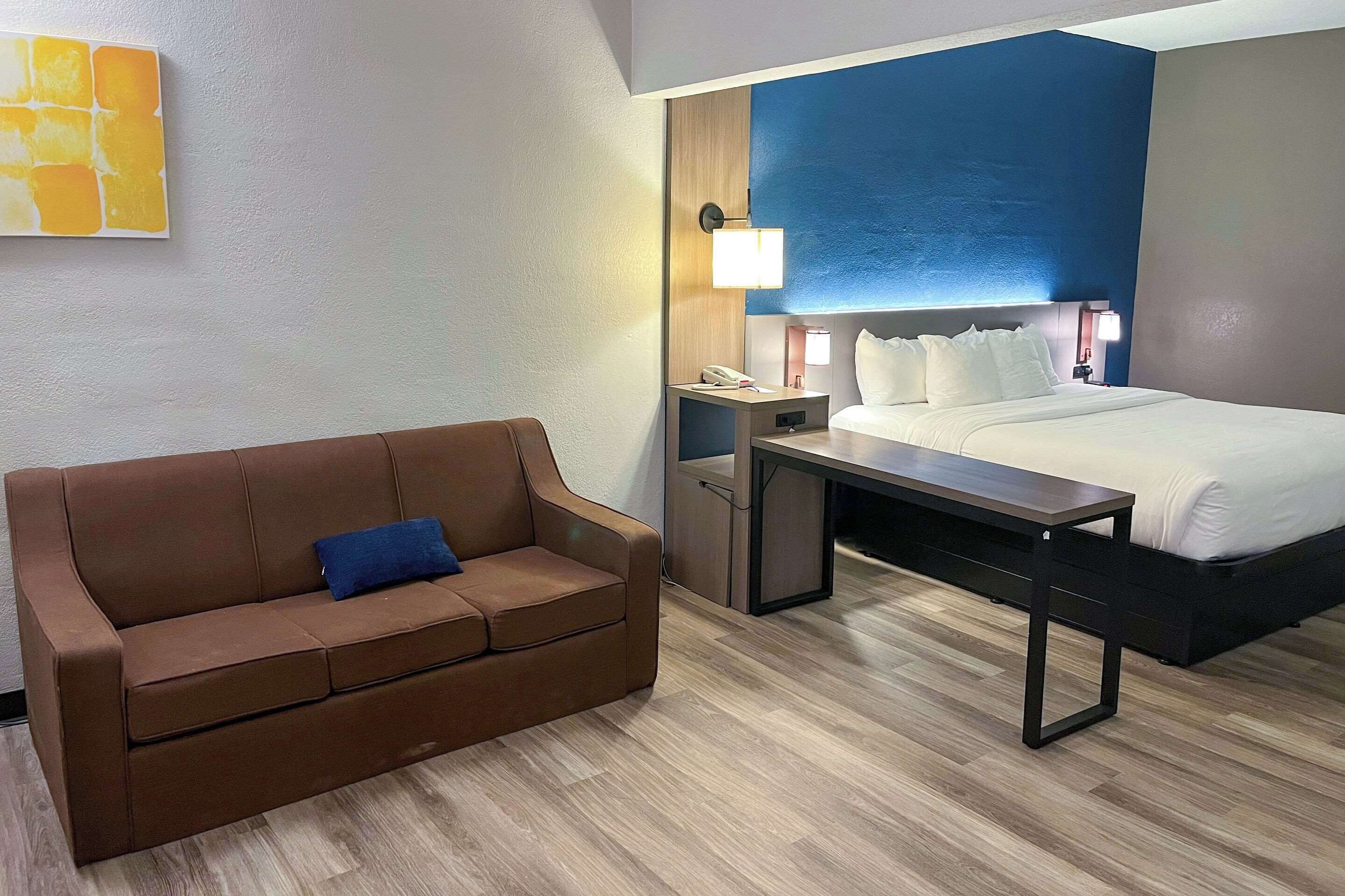 Una habitación de hotel con una cama, una mesita de noche, un sofá marrón y una obra de arte en la pared.