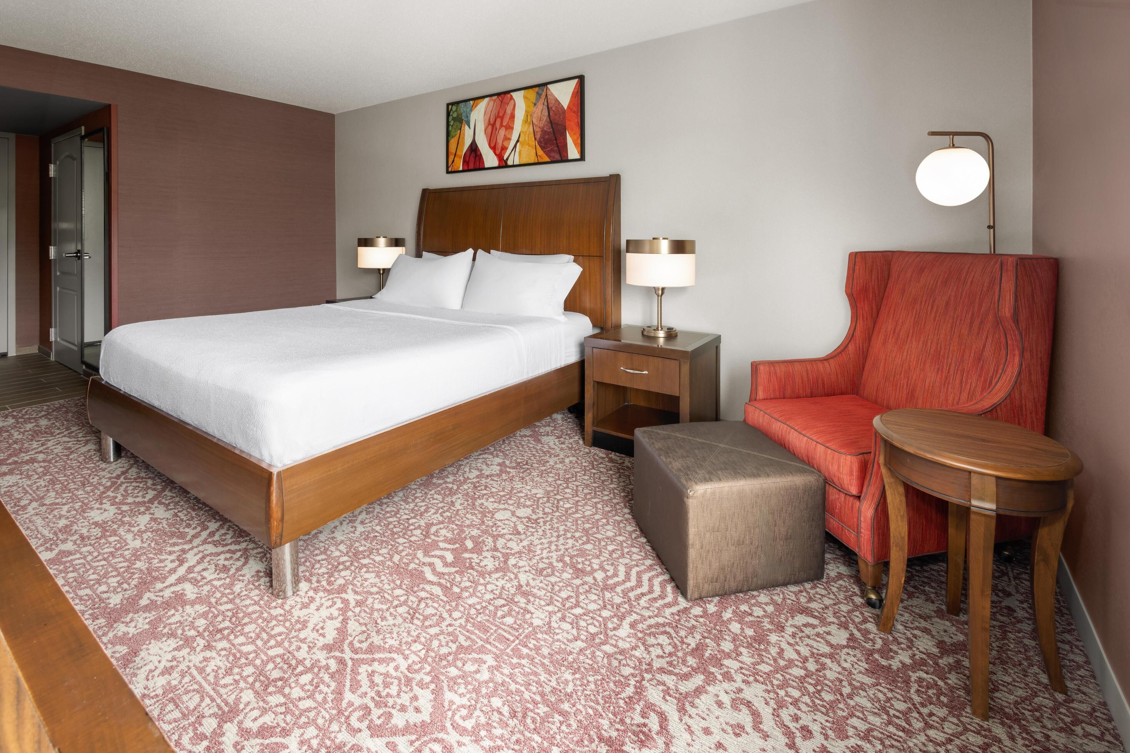 Una habitación de hotel con una cama grande, una mesita de noche de madera, un sillón rojo, una pequeña mesa y una lámpara fijada a la pared.