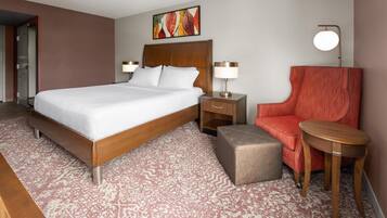 Una habitación de hotel con una cama grande, una mesita de noche de madera, un sillón rojo, una pequeña mesa y una lámpara fijada a la pared.