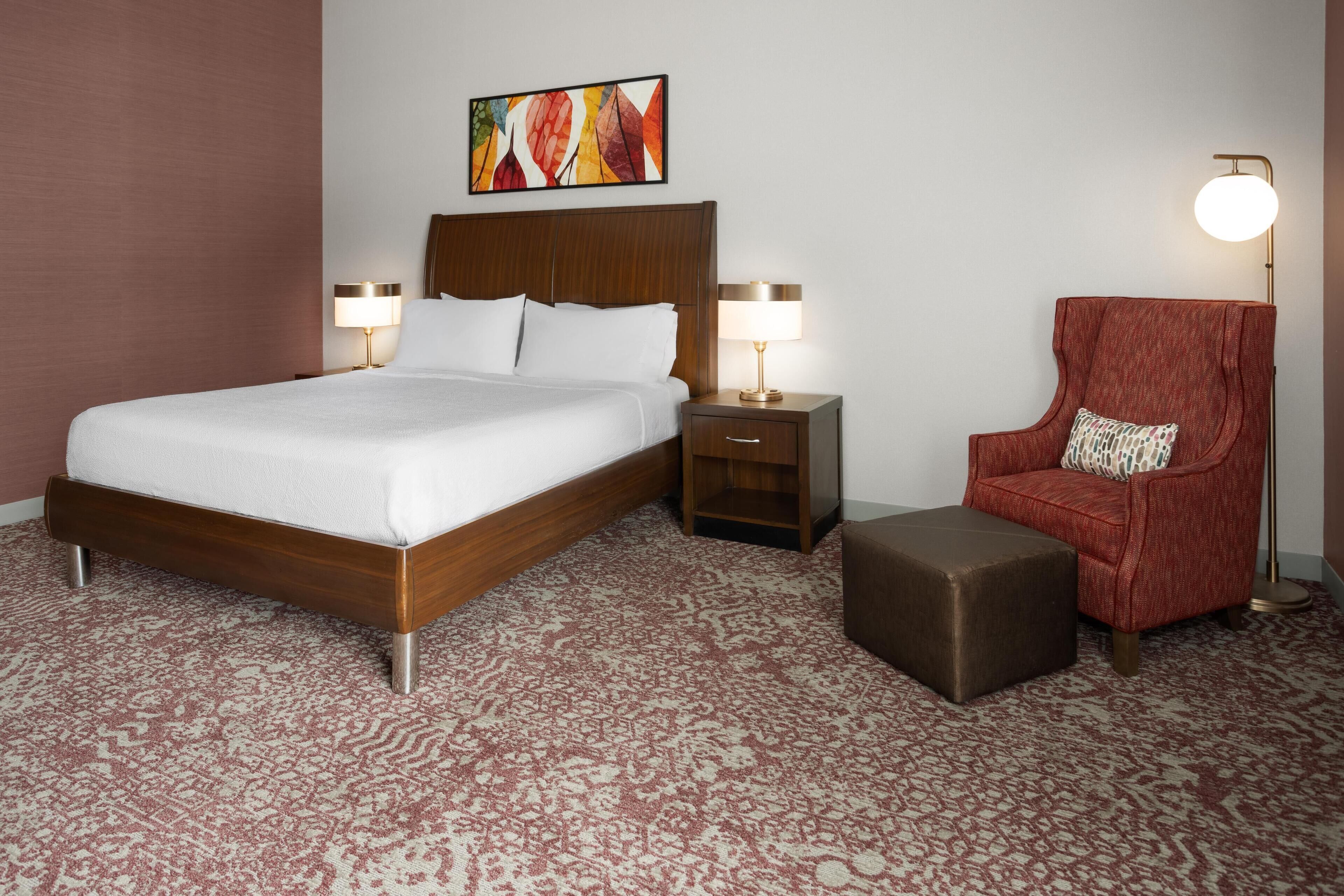 A hotel room with a large bed, two bedside tables with lamps, a red armchair, and a small ottoman.
