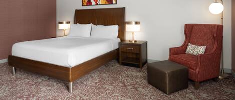 A hotel room with a large bed, two bedside tables with lamps, a red armchair, and a small ottoman.