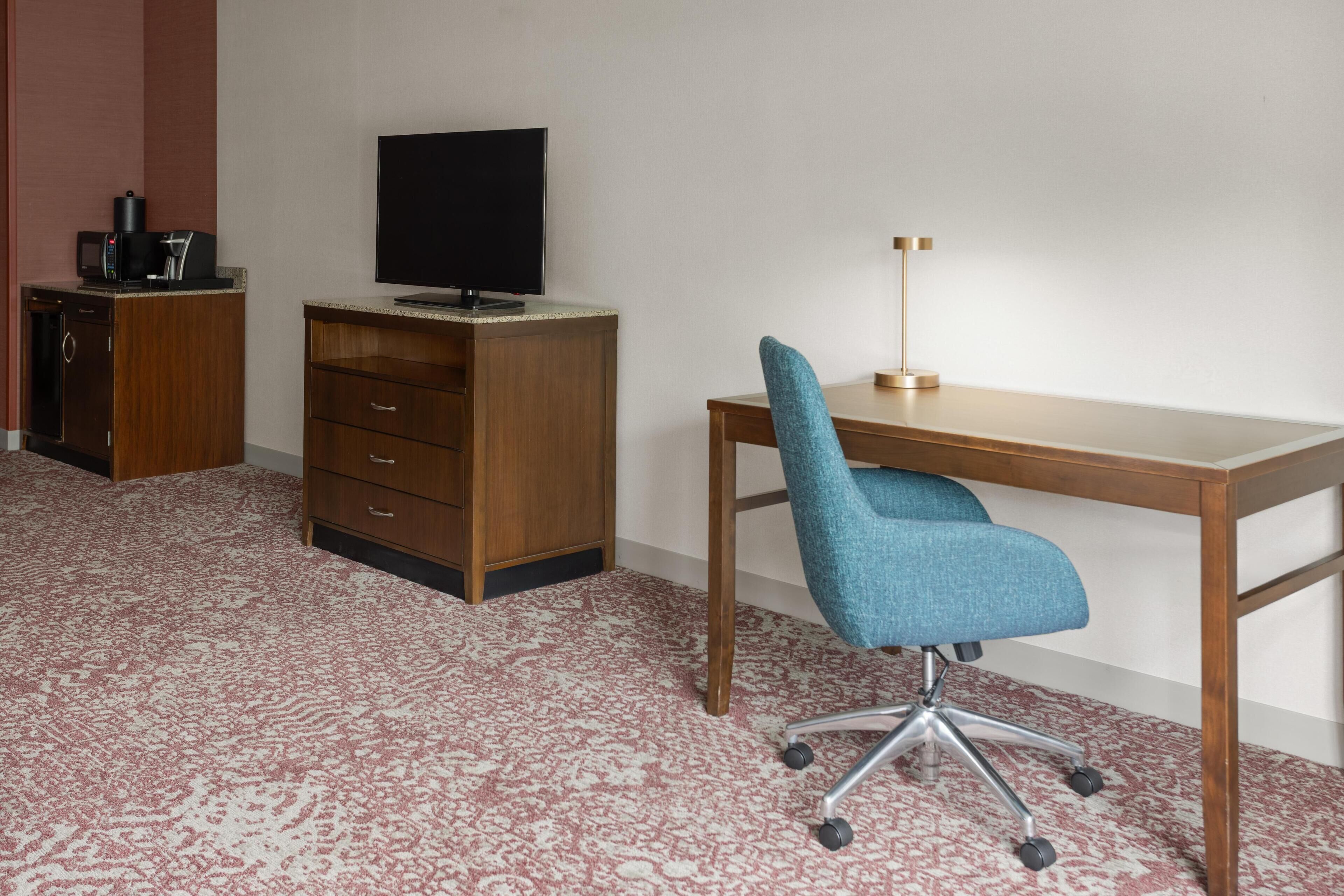 A room with a wooden desk, a blue office chair, a television on a cabinet, and a coffee maker.