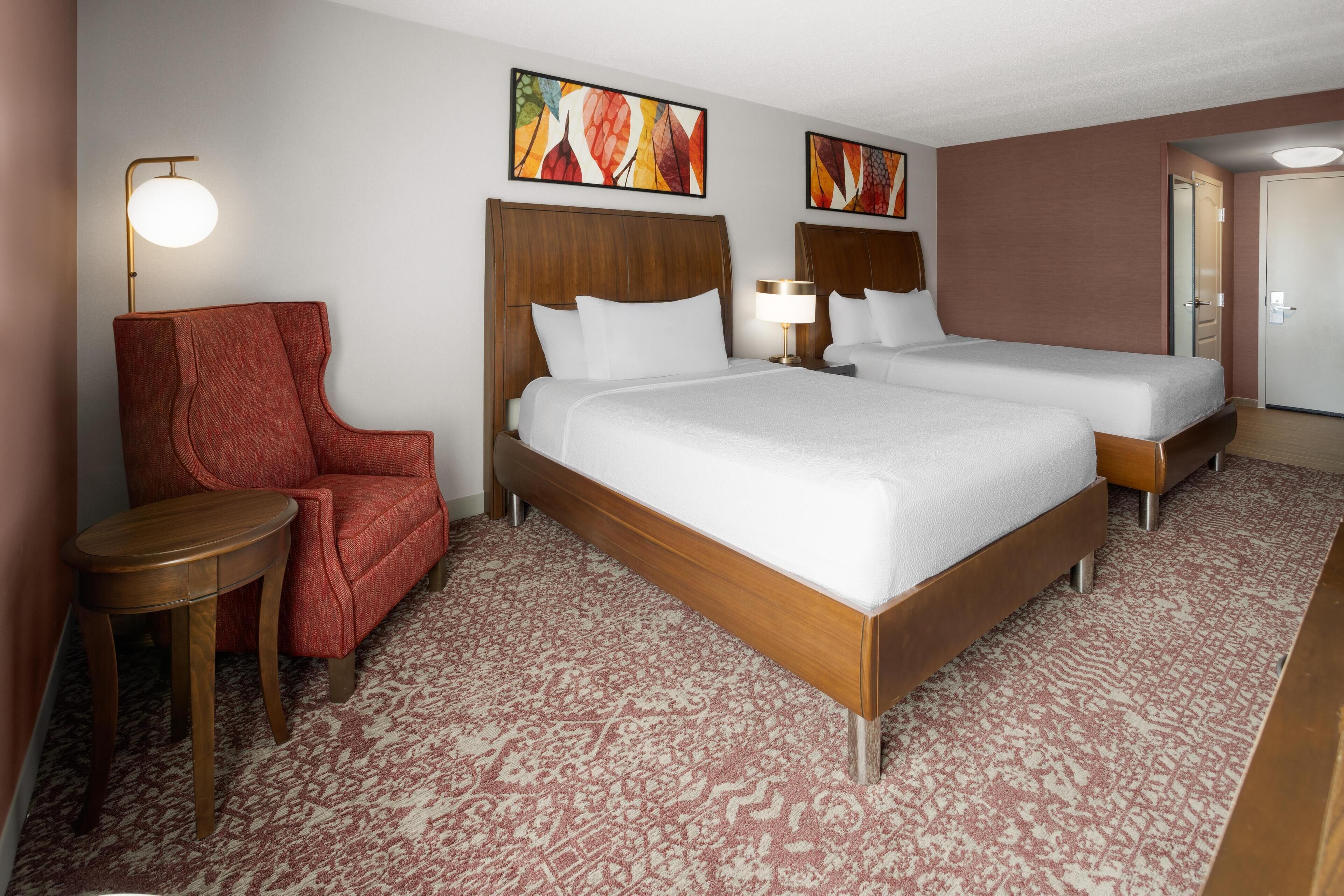 Habitación de hotel con dos camas, un sillón rojo, una mesita y dos lámparas de pared.