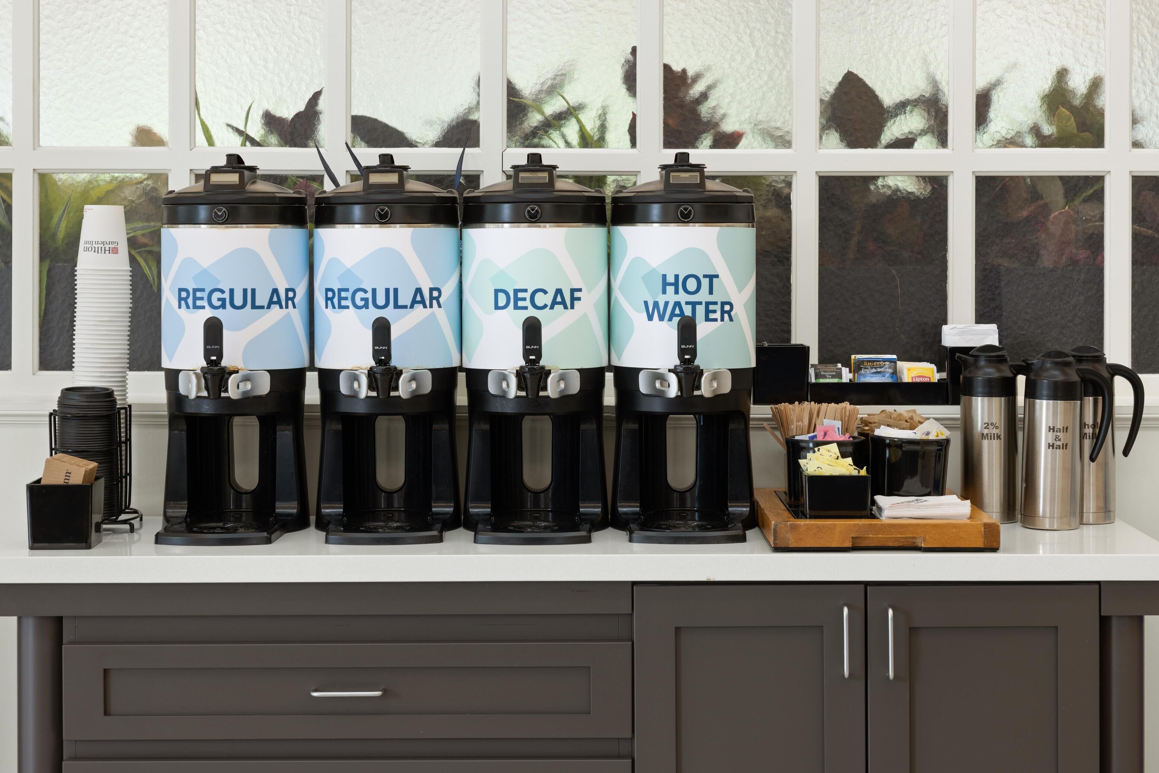 Un coin café est aménagé avec quatre distributeurs étiquetés « Café normal », « Café normal », « Café décaféiné » et « Eau chaude ». On y trouve des tasses à café, un thermos et un plateau avec des ustensiles.