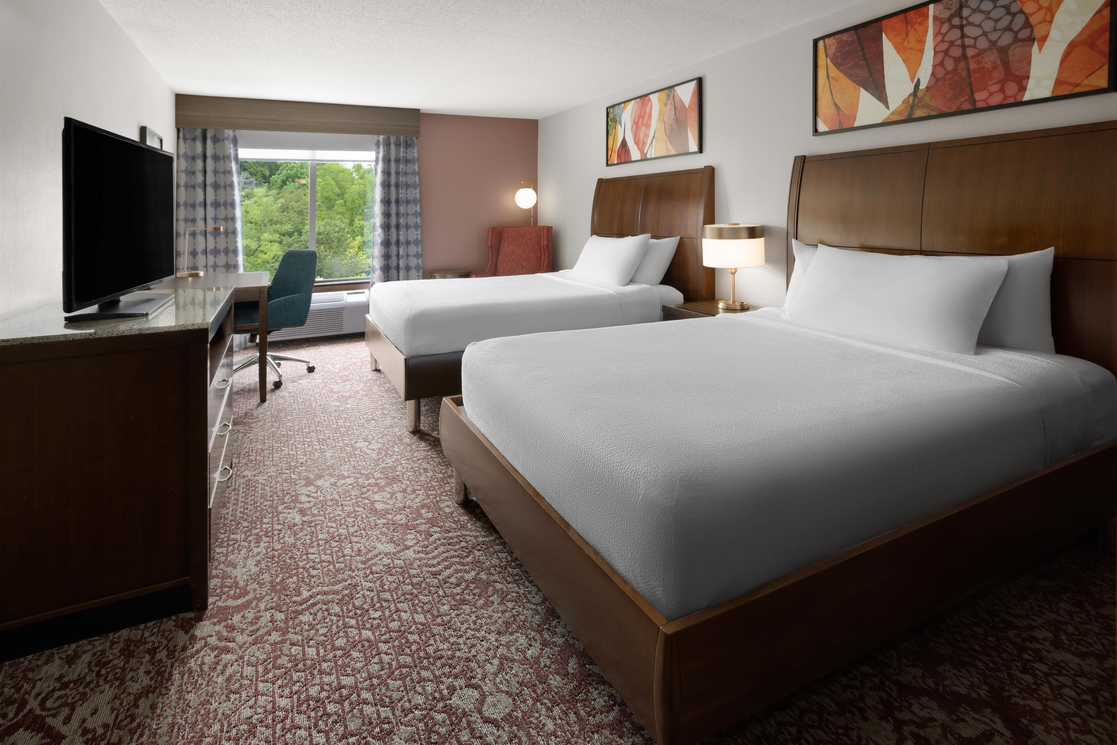 A hotel room with two beds, a desk with a TV, a chair, and a large window with curtains.