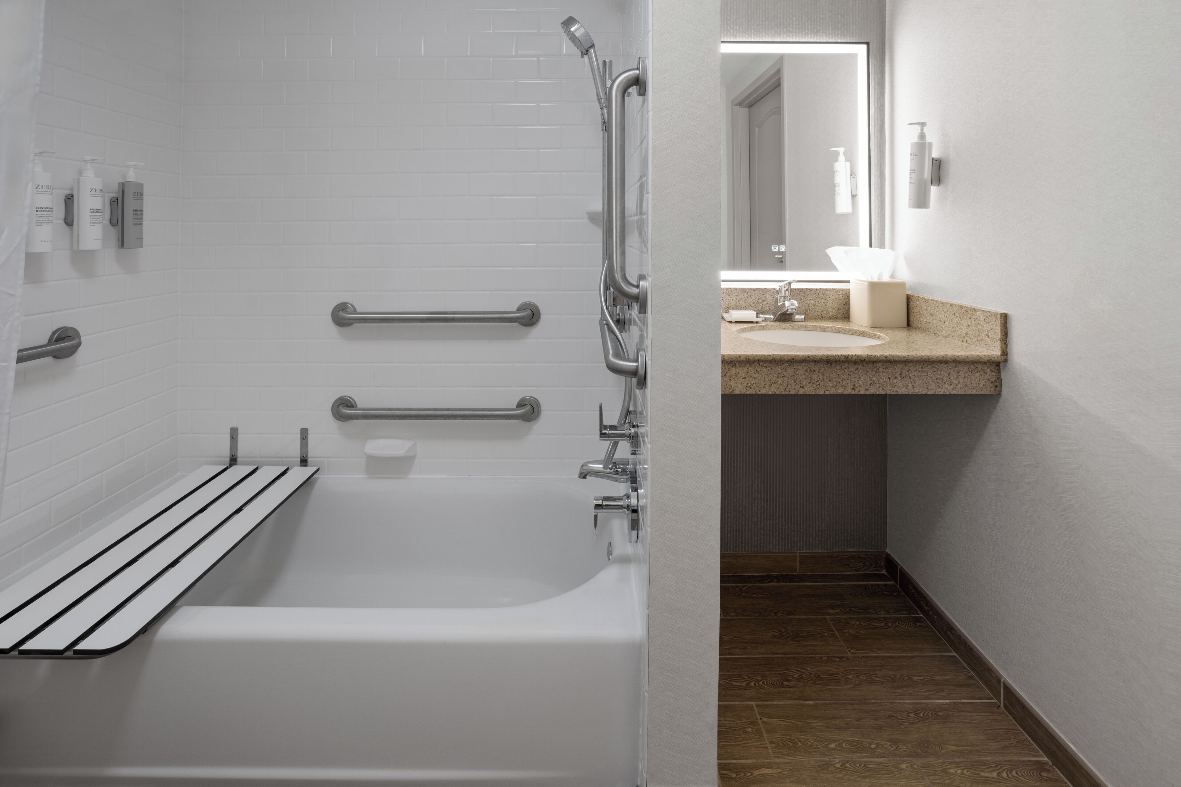 A modern bathroom with a white bathtub, grab bars, a shower with a handheld showerhead, a sink with a countertop, and a mirror above the sink.