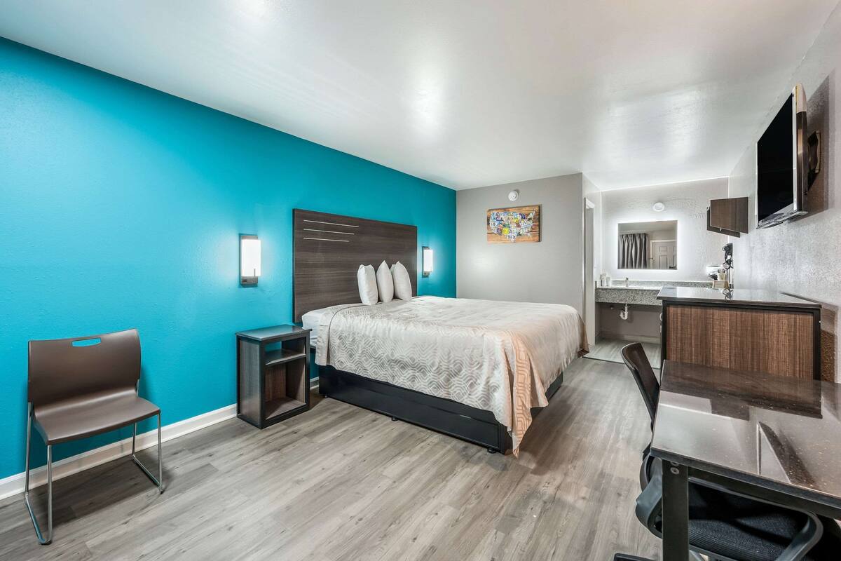 A modern hotel room with a teal accent wall, a bed with a headboard, a desk with a chair, a small table, and a built-in TV.