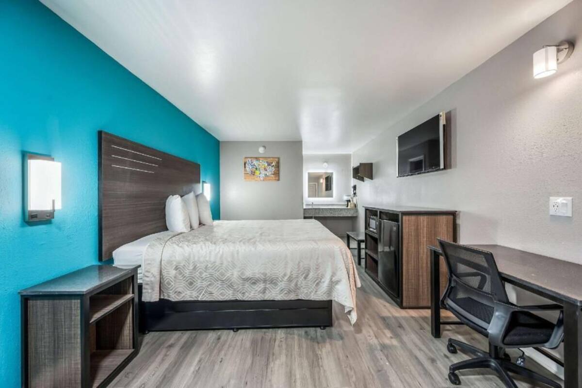 A modern hotel room with a bed, desk, and kitchenette.