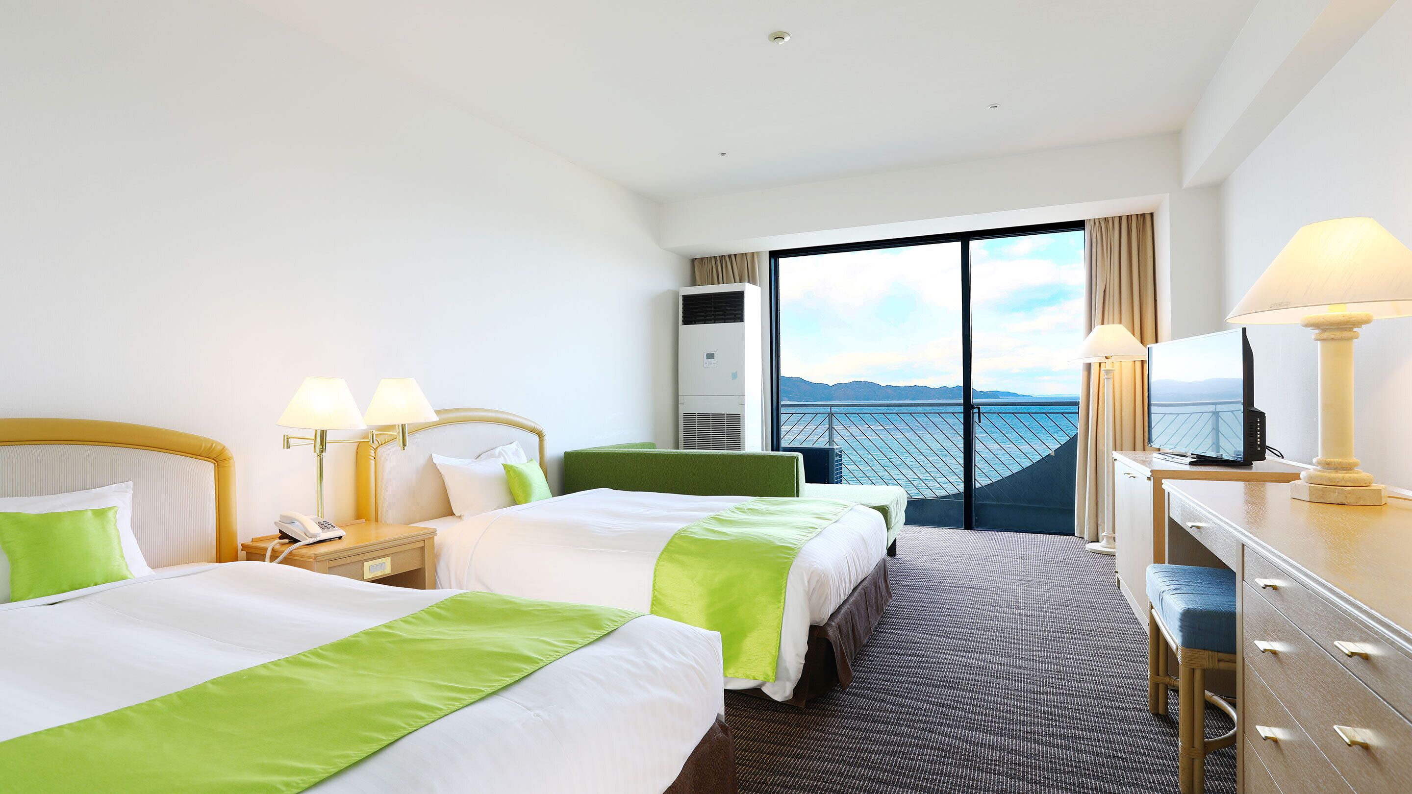 A hotel room with two beds, a desk, a TV, and a view of the sea.