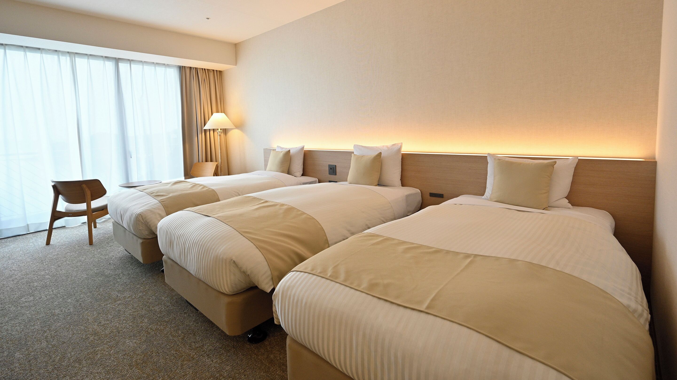 A hotel room with two beds, a chair, a lamp, and a large window with curtains.