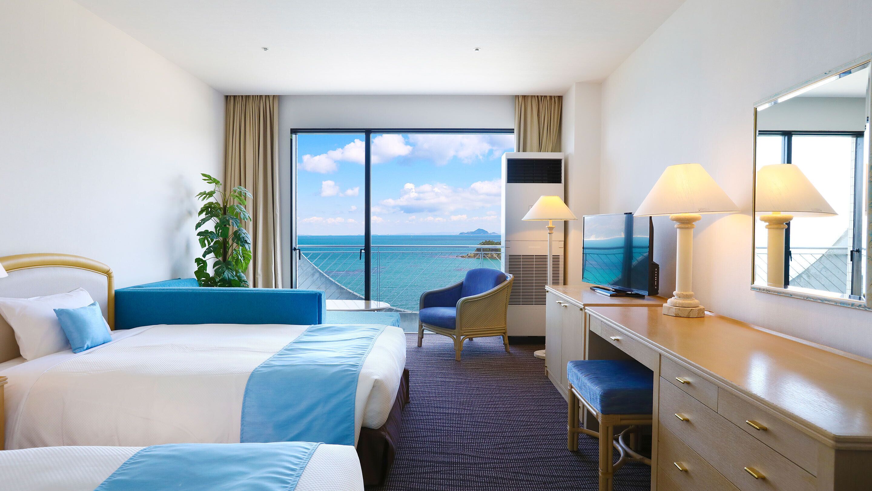 A hotel room with two beds, a desk with a TV, a chair, and a large window with a view of the sea.