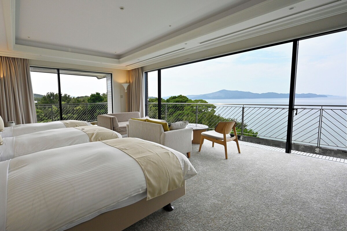 A hotel room with two beds, a balcony with a view of the sea, and a dining area.