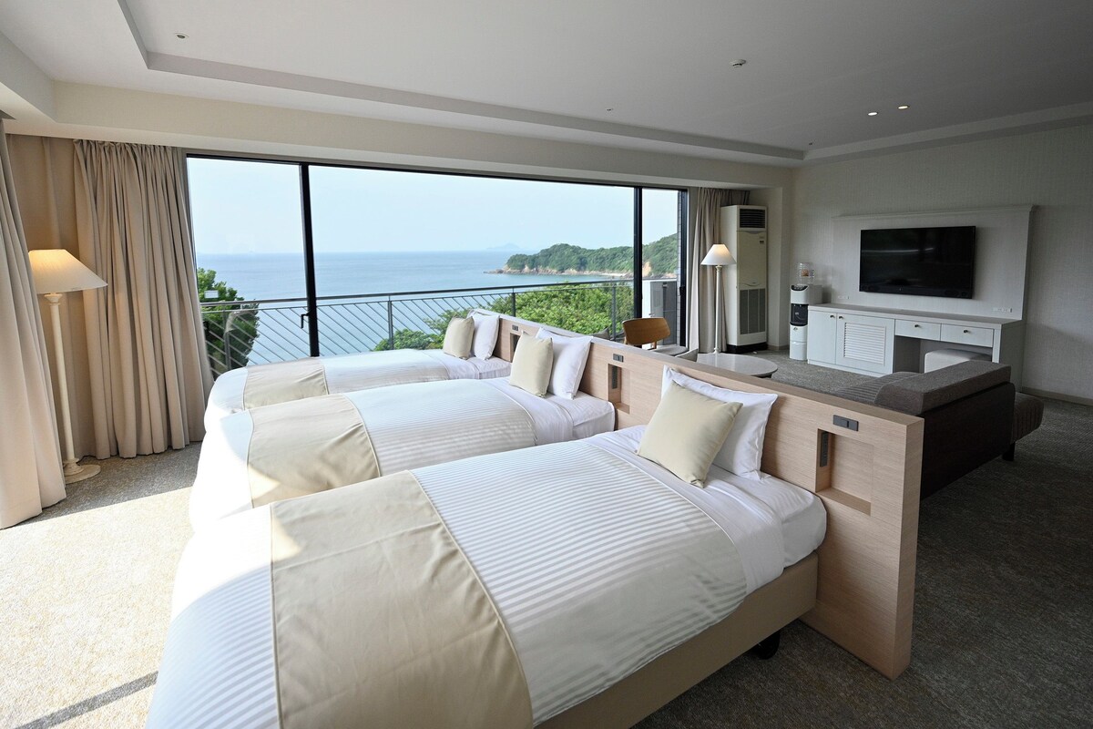 A hotel room with two beds, a TV, and a balcony with a view of the sea.