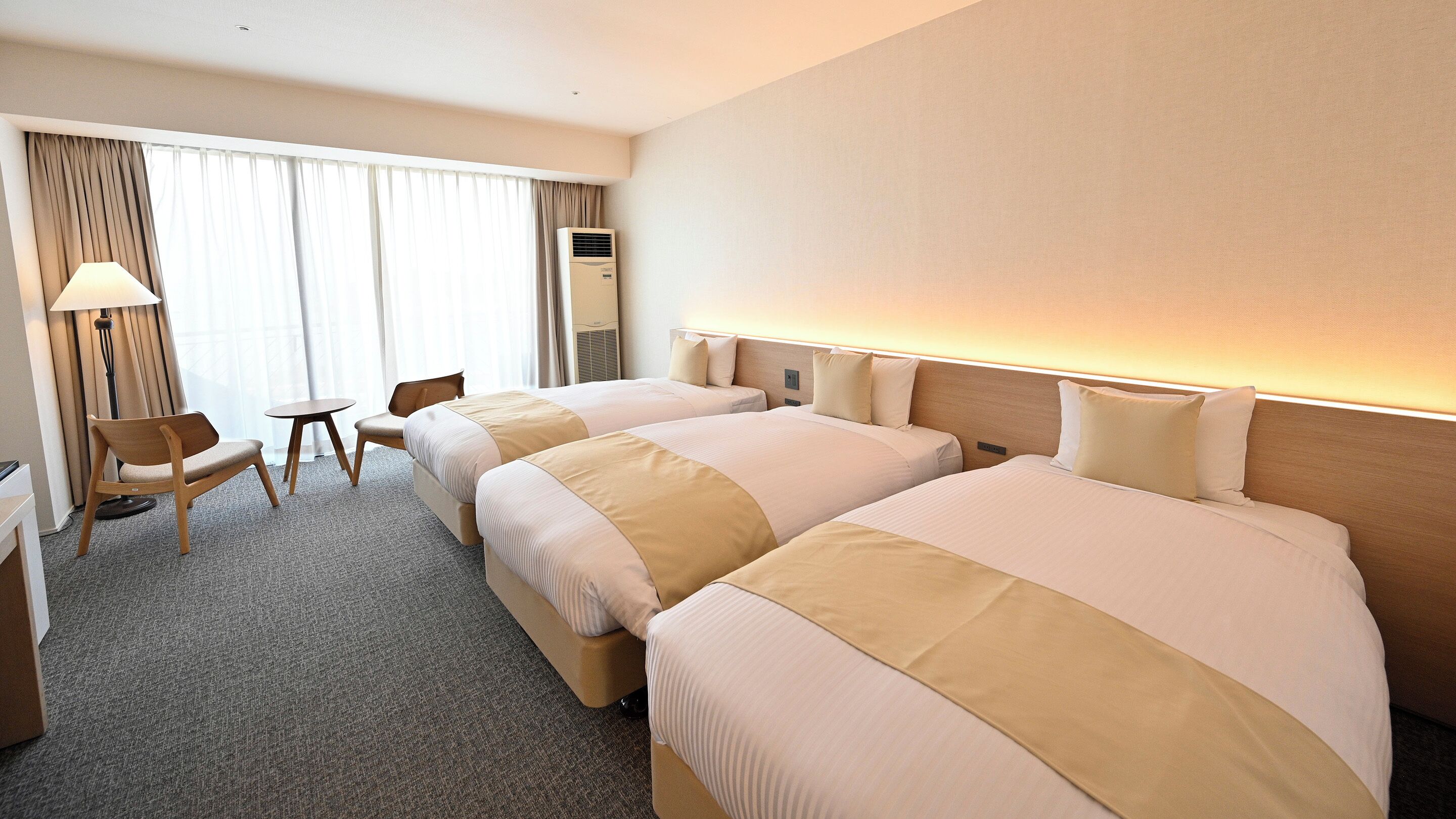A hotel room with two beds, a desk, a chair, and a lamp.