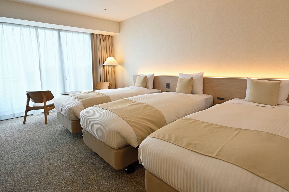 A hotel room with two beds, a chair, a lamp, and a large window with curtains.