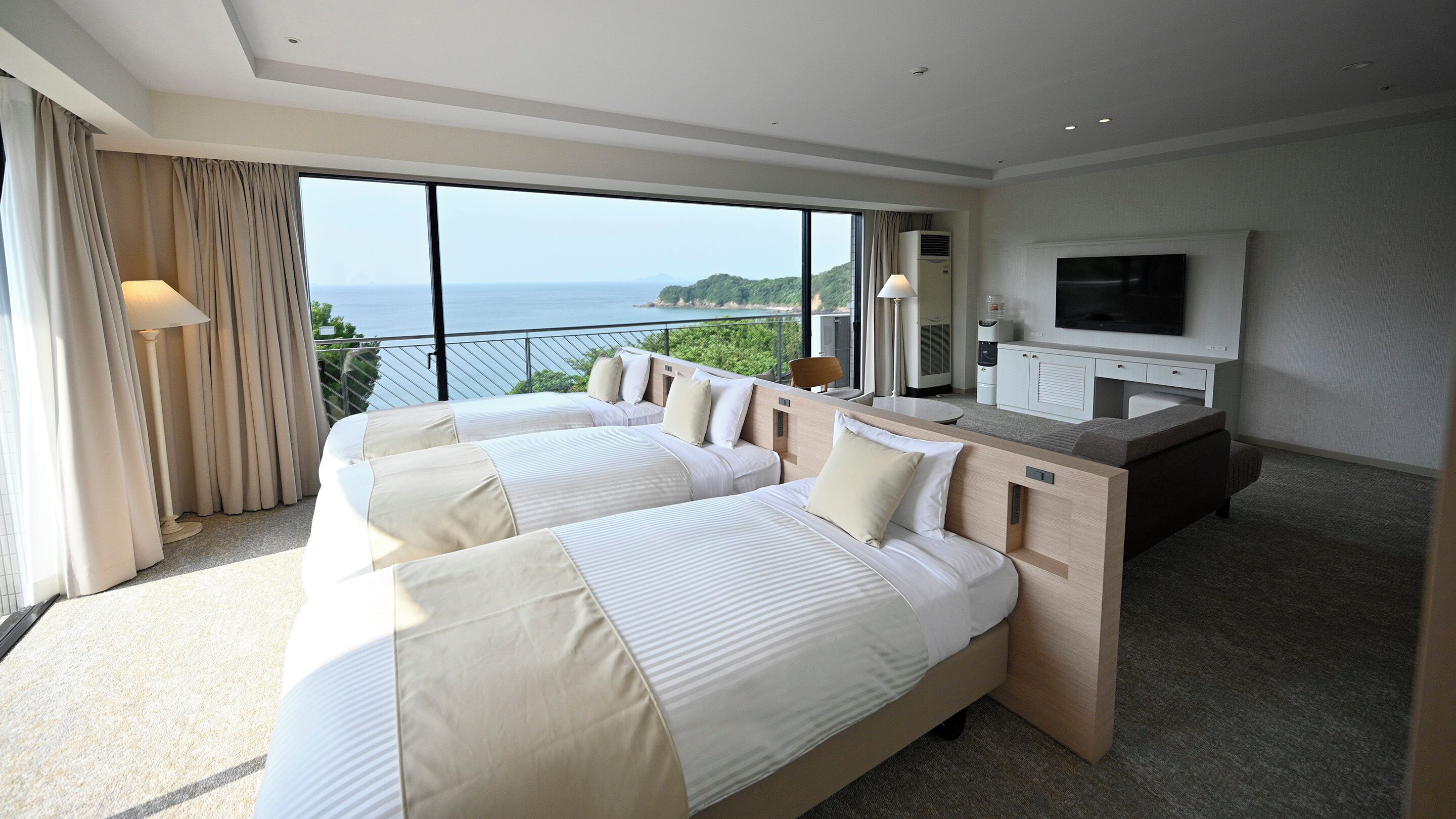 A hotel room with two beds, a TV, and a balcony with a view of the sea.