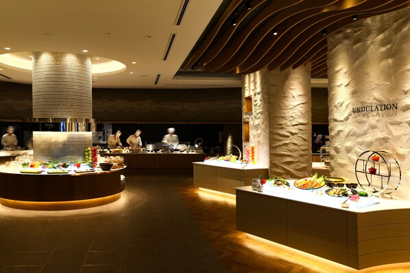 Un hall d’hôtel moderne avec un comptoir d’accueil central, une colonne décorative et un espace buffet proposant divers plats.