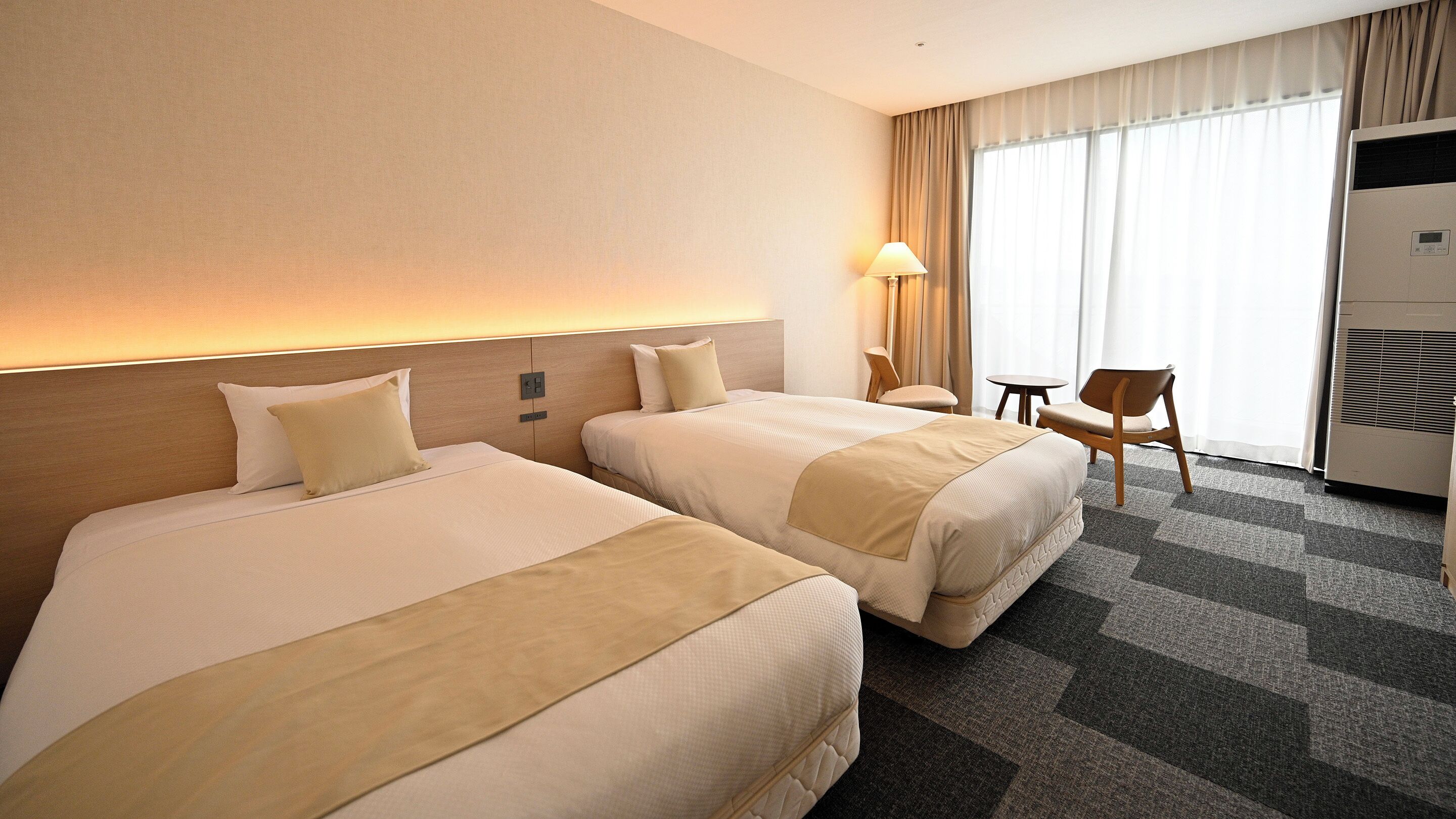 A hotel room with two beds, a desk, a chair, and a large window with curtains.