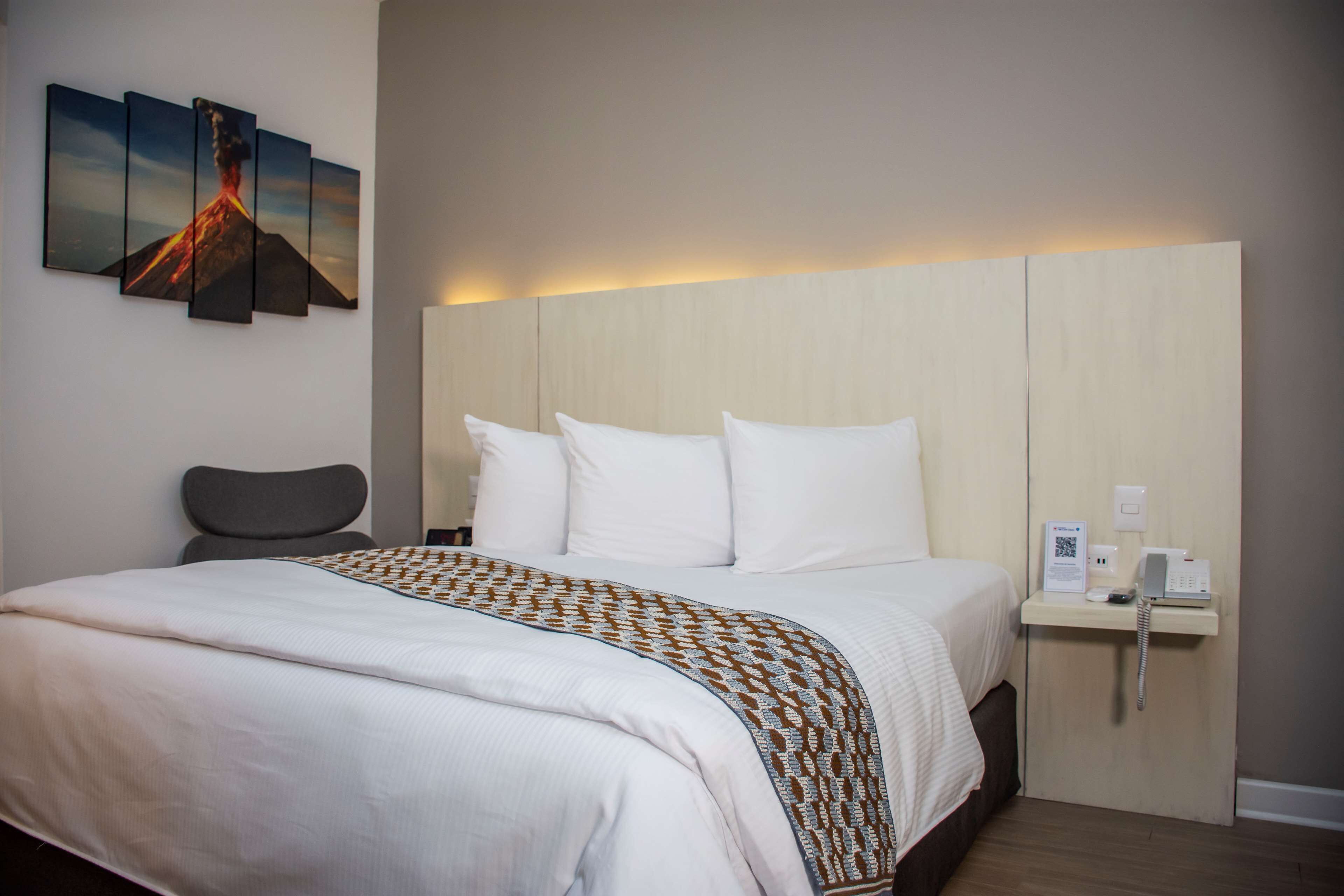 Habitación de hotel con una cama grande, una mesita de noche con un teléfono y una pequeña señal, y una obra de arte colgada en la pared que representa un volcán.