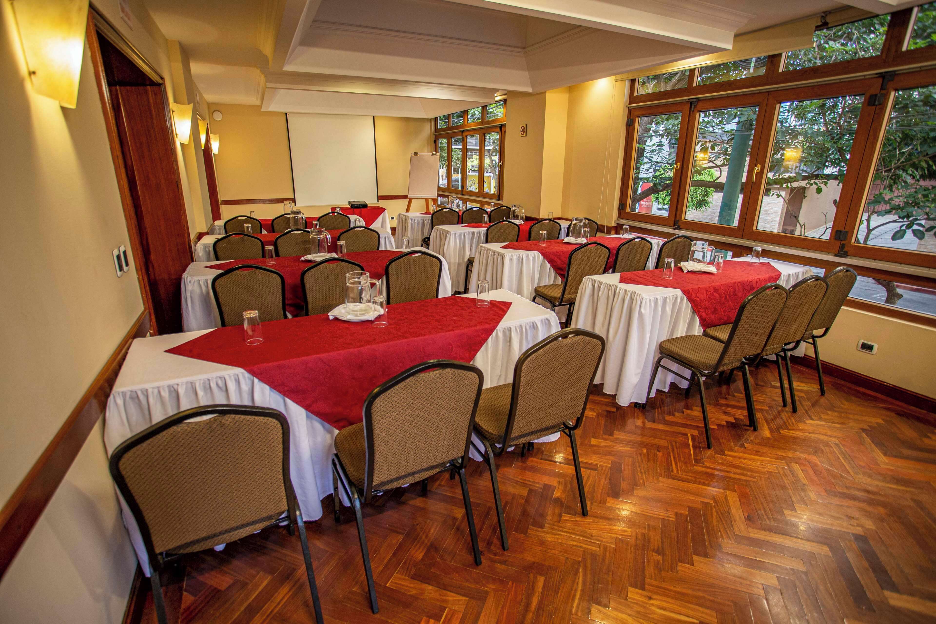 Un salón de conferencias con varias mesas y sillas, ventanas amplias que dan a zonas verdes, y un suelo de madera en patrón espiga.
