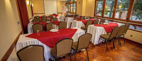 Un salón de conferencias con varias mesas y sillas, ventanas amplias que dan a zonas verdes, y un suelo de madera en patrón espiga.