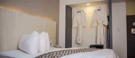 A hotel room with a bed, pillows, a bedside table with a phone, and two robes hanging on hooks.