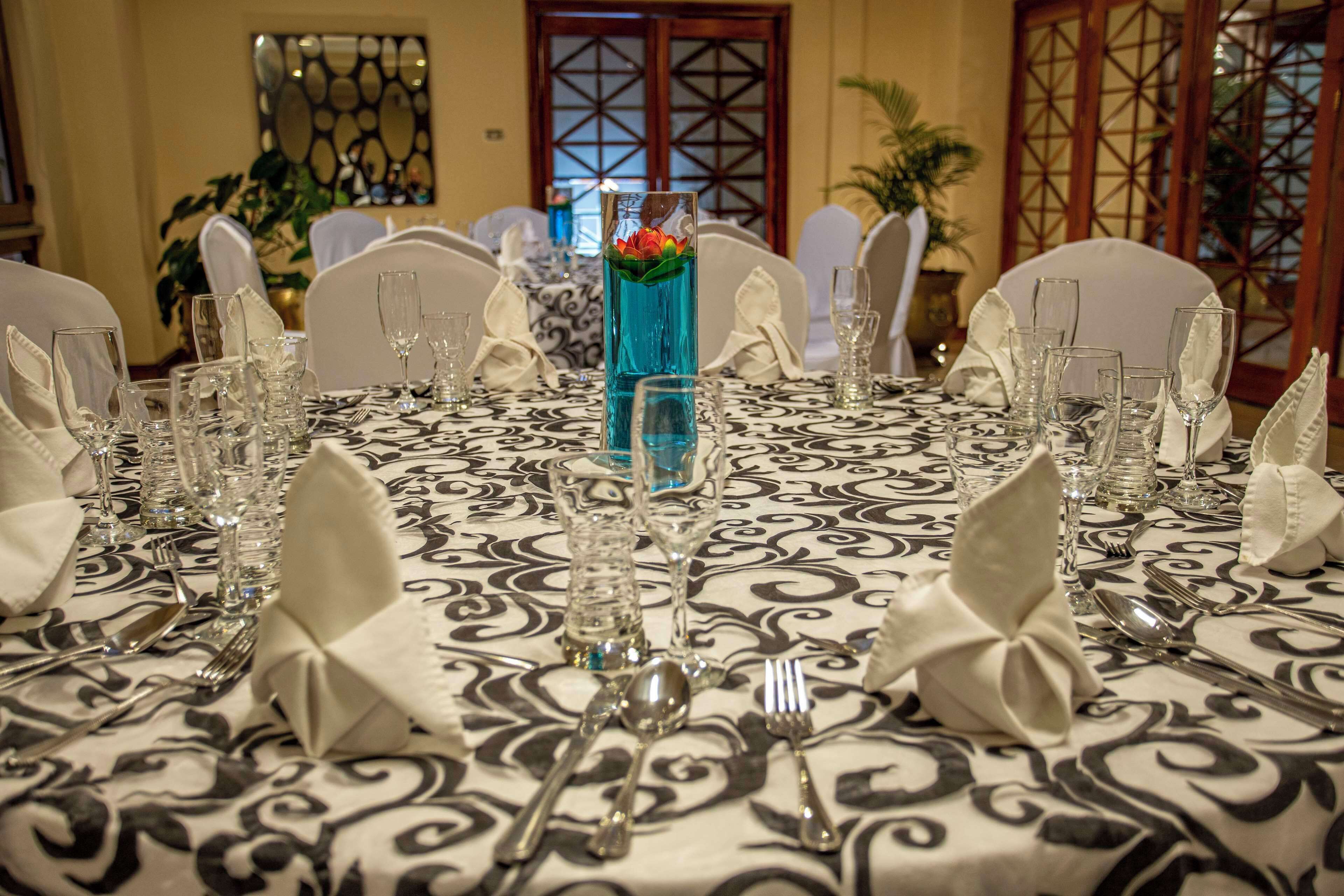 A dining table set for a formal meal with a patterned tablecloth, white chairs, and a centerpiece with a blue vase and flowers.