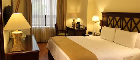 A hotel room with a large bed, two bedside tables with lamps, a desk with a chair, and a window with curtains.