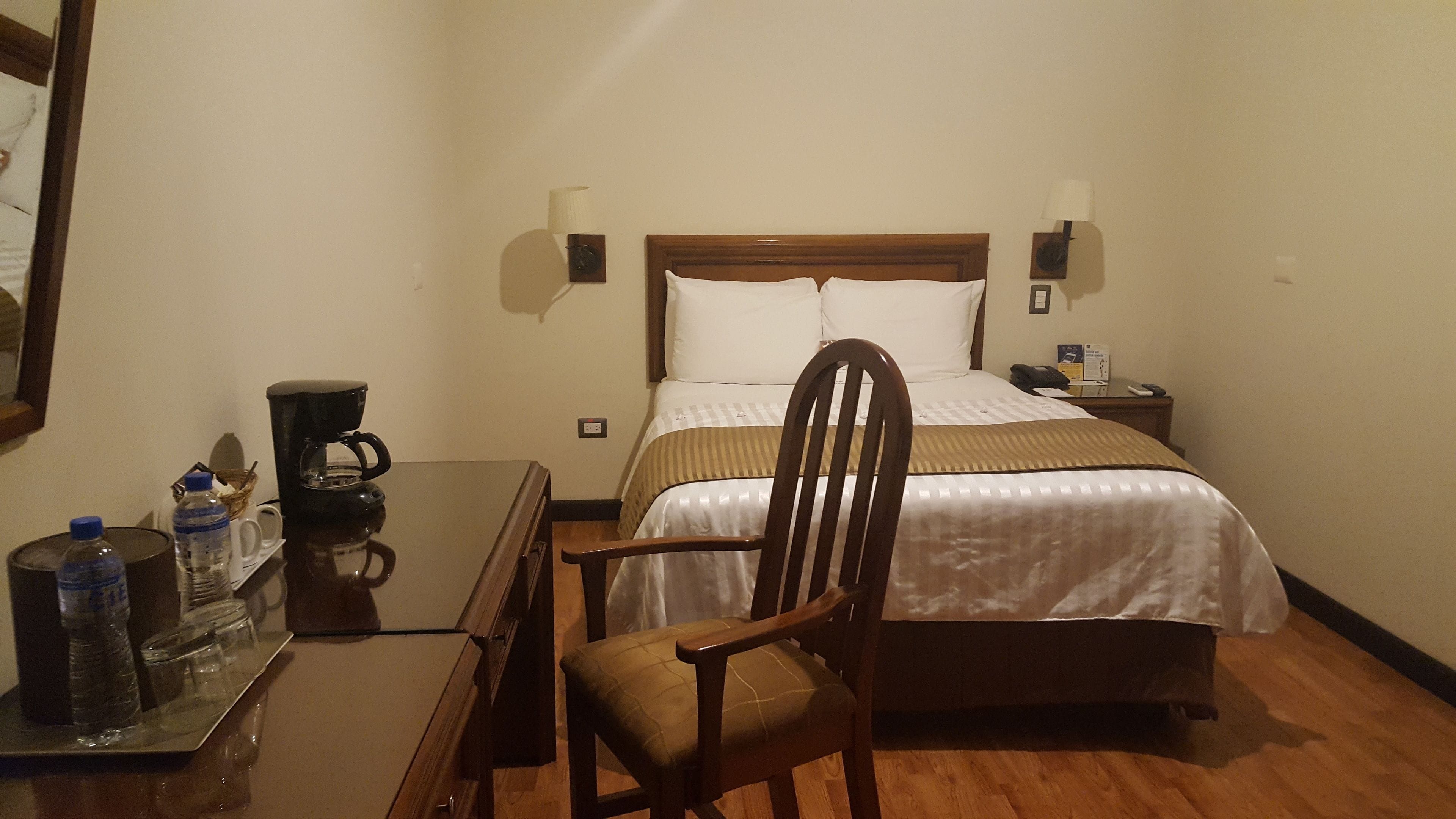 Une chambre d’hôtel avec un lit, une chaise en bois, une machine à café et une table avec des bouteilles d’eau et des verres.