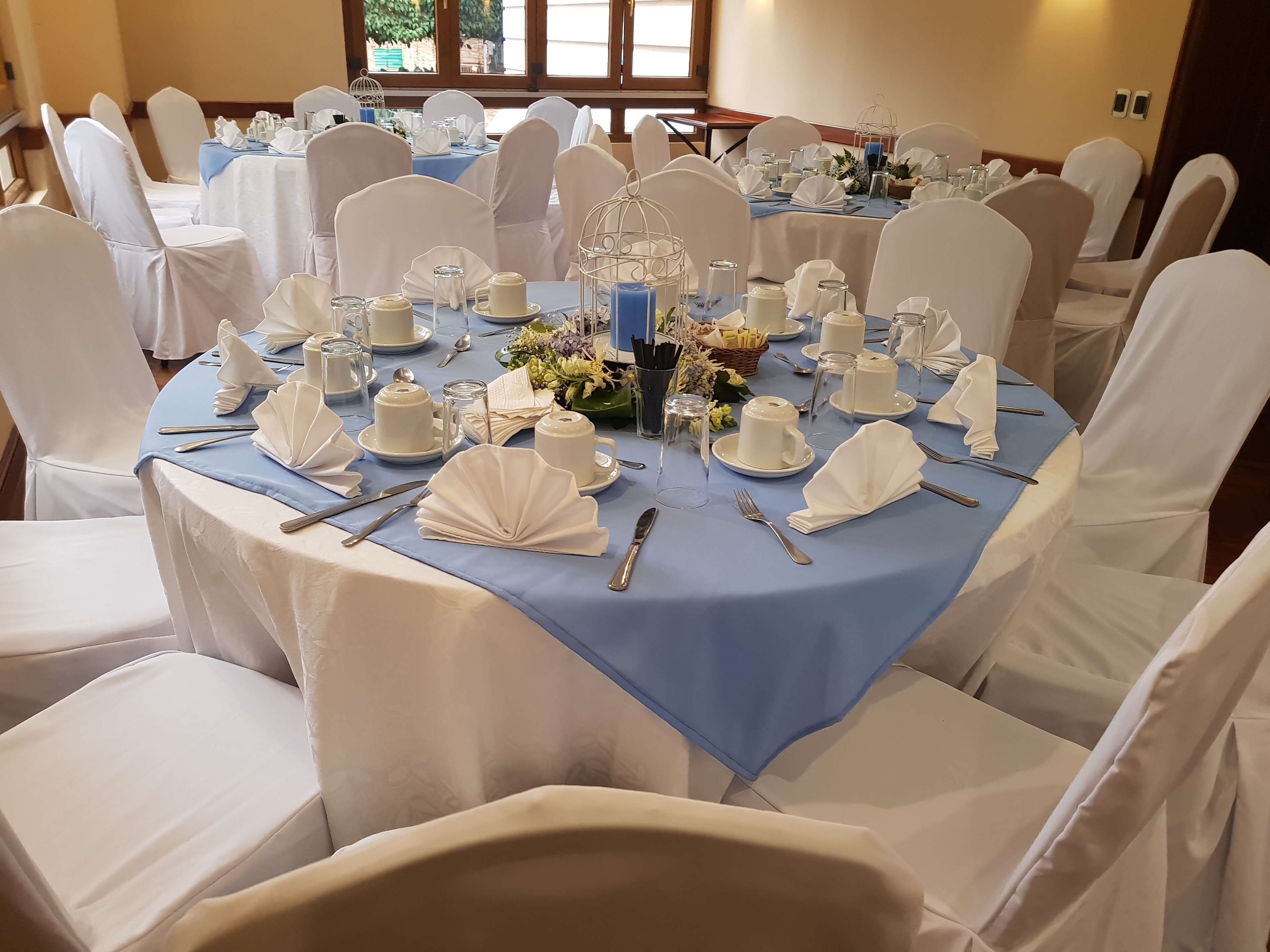 A banquet hall with round tables covered in blue tablecloths, white chairs, and white table settings including plates, cups, and cutlery.
