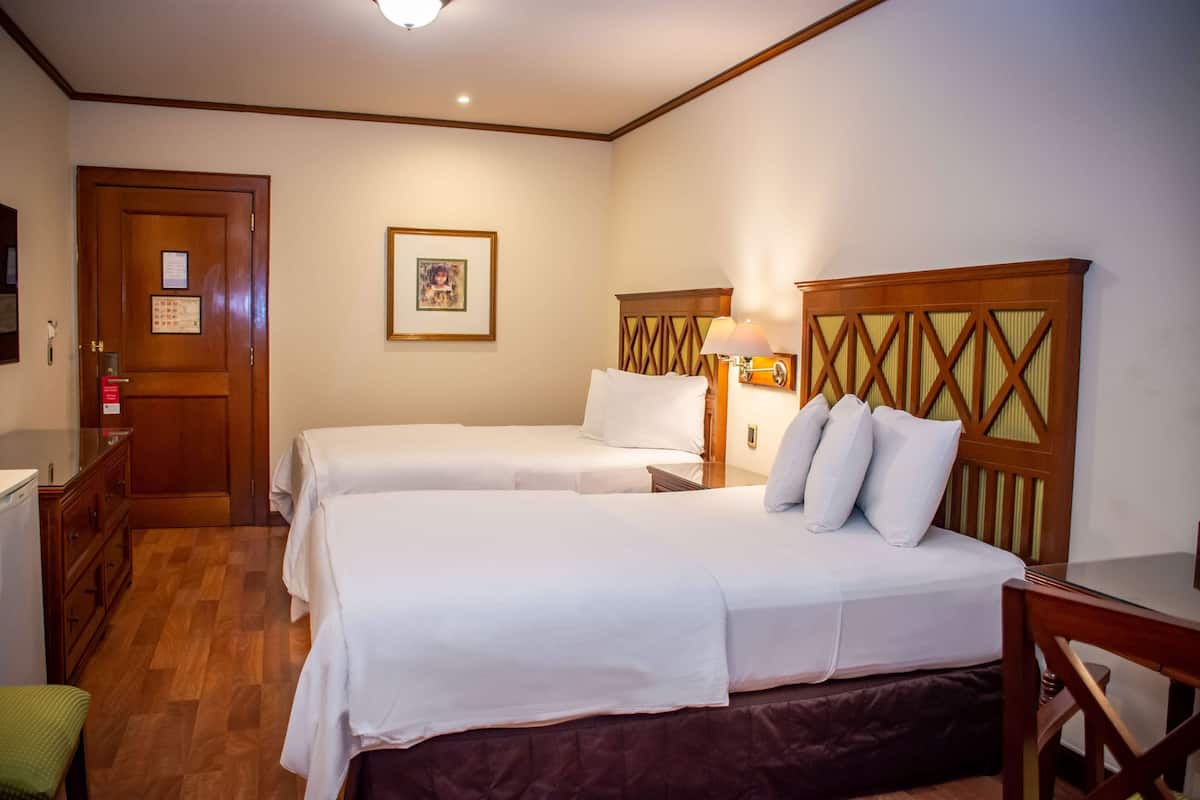 Een hotelkamer met twee bedden, een houten hoofdbord, een nachtkastje met een lamp, een dressoir en een ingelijst schilderij aan de muur.