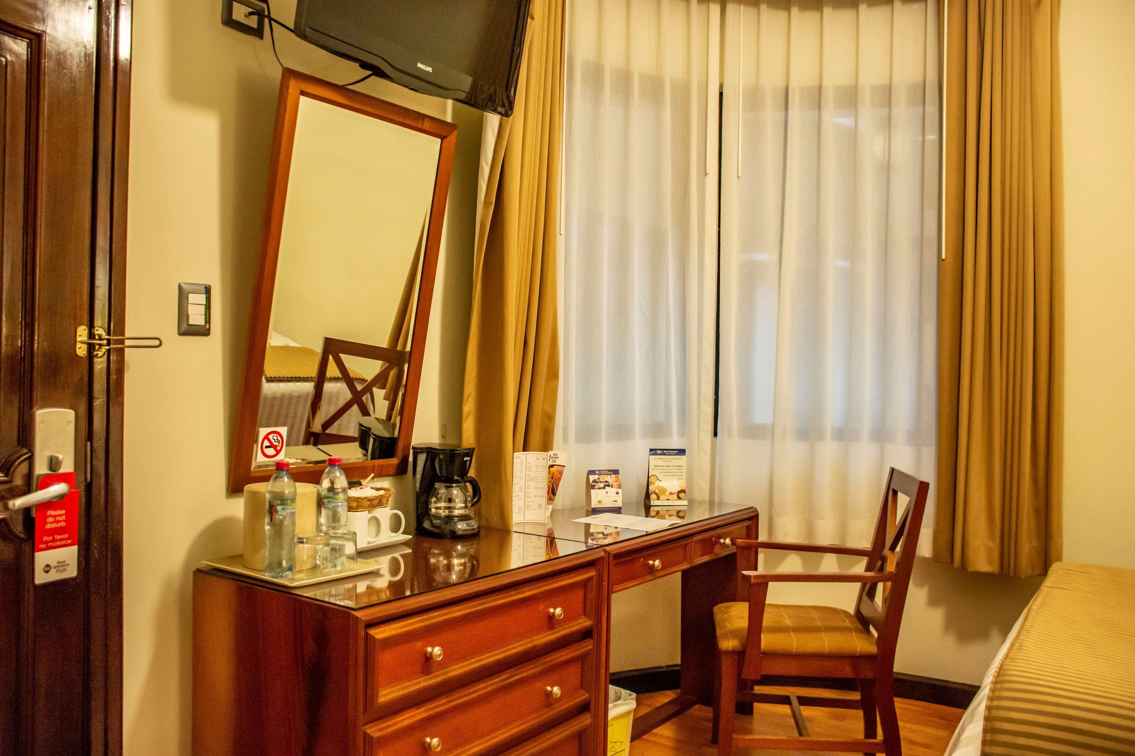 Habitación de hotel con escritorio de madera, una silla, cama, televisor y ventana con cortinas.