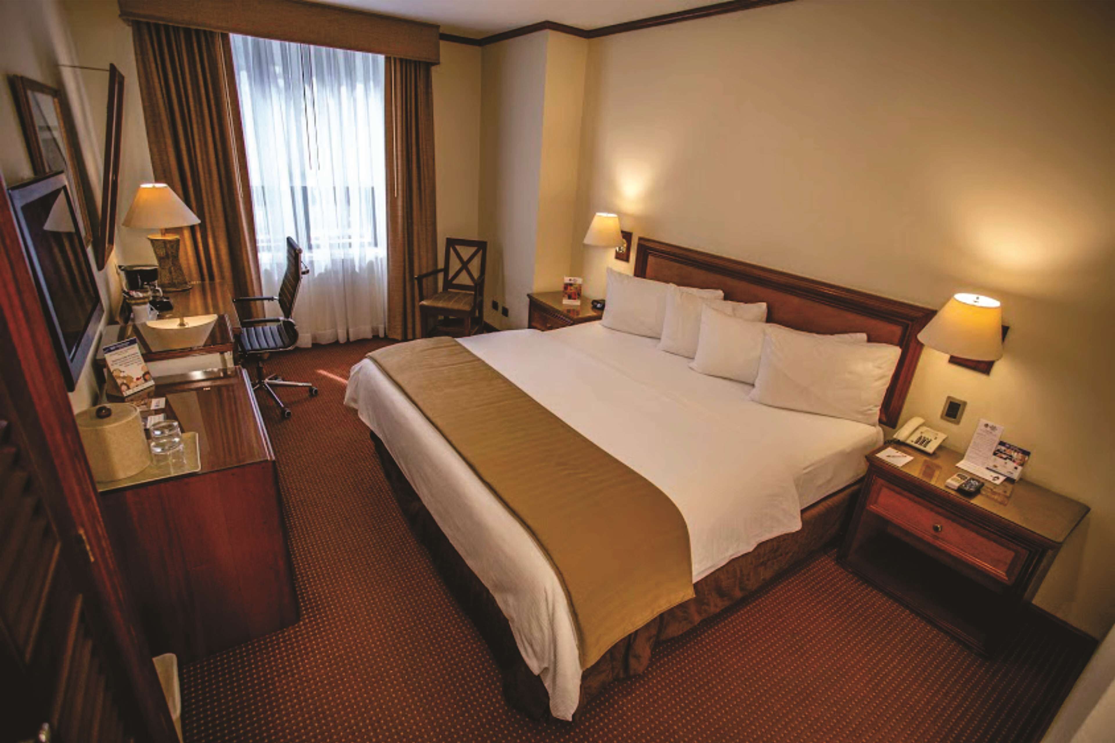 A hotel room with a large bed, two bedside tables with lamps, a desk with a chair, a chair by the window, and a mirror on the wall.