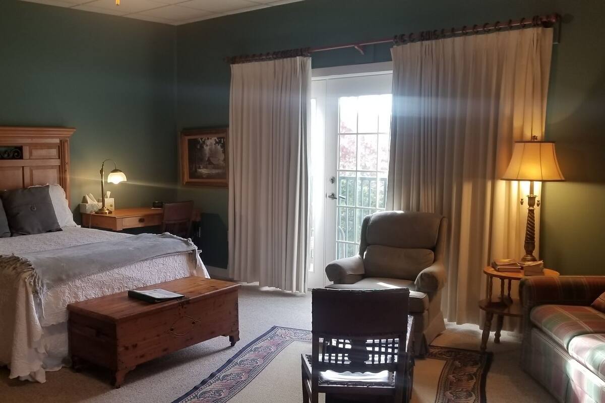 A well-lit bedroom with a large bed, a wooden nightstand, a chair, a sofa, and a desk.