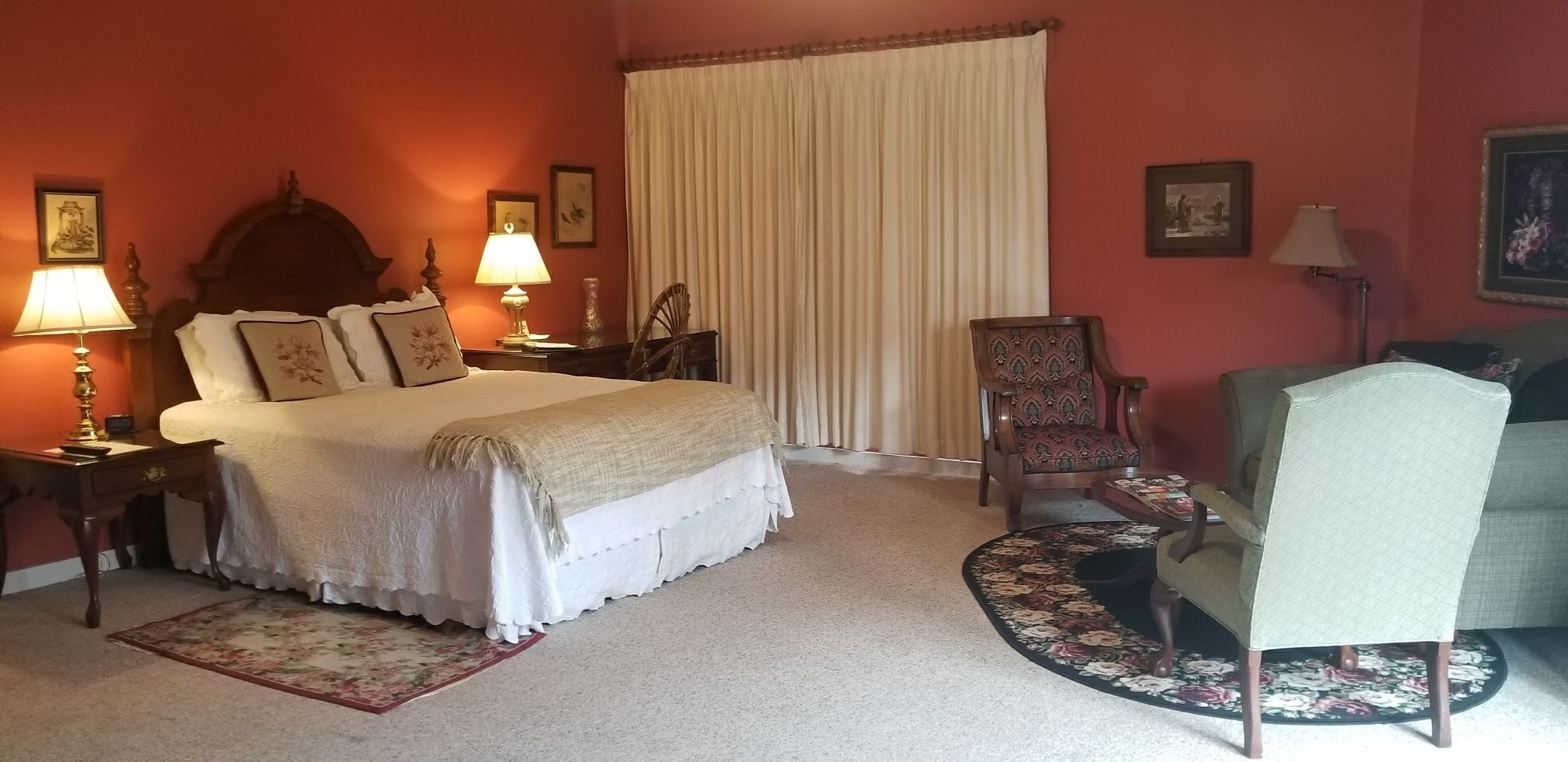Un dormitorio con una cama, dos mesitas de noche con lámparas, una silla, un sofá, un ventanal con cortinas y suelo alfombrado.