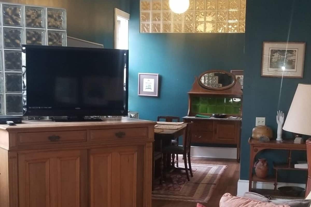 A living room with a television, wooden cabinets, a sofa with patterned cushions, and a kitchen area in the background.